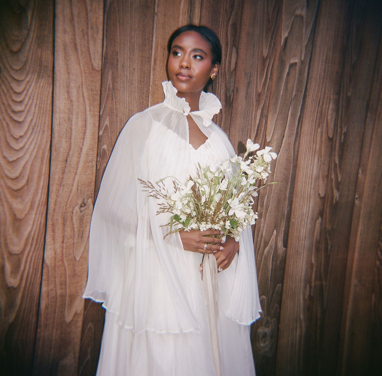A Black bride in a flowing white chiffon gown with cape holds a white wildflower bouquet against a wooden backdrop.