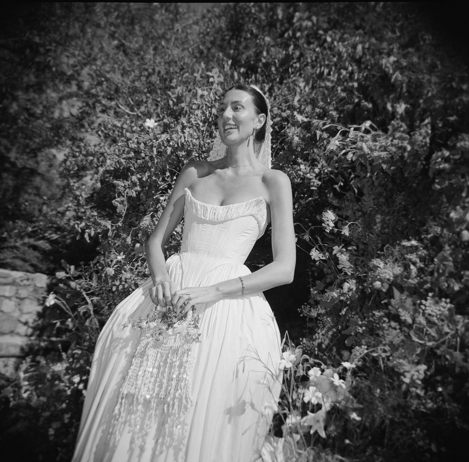 Black and white photo of a bride in a flowing gown holding a bouquet, standing among lush greenery.