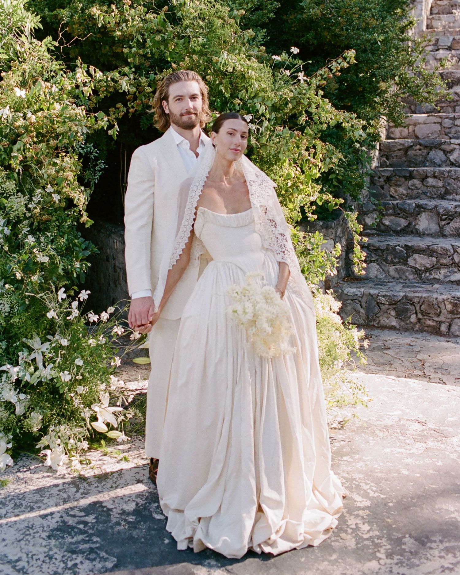 Bride in ivory gown with veil and groom in white suit pose together outdoors near stone wall and greenery.