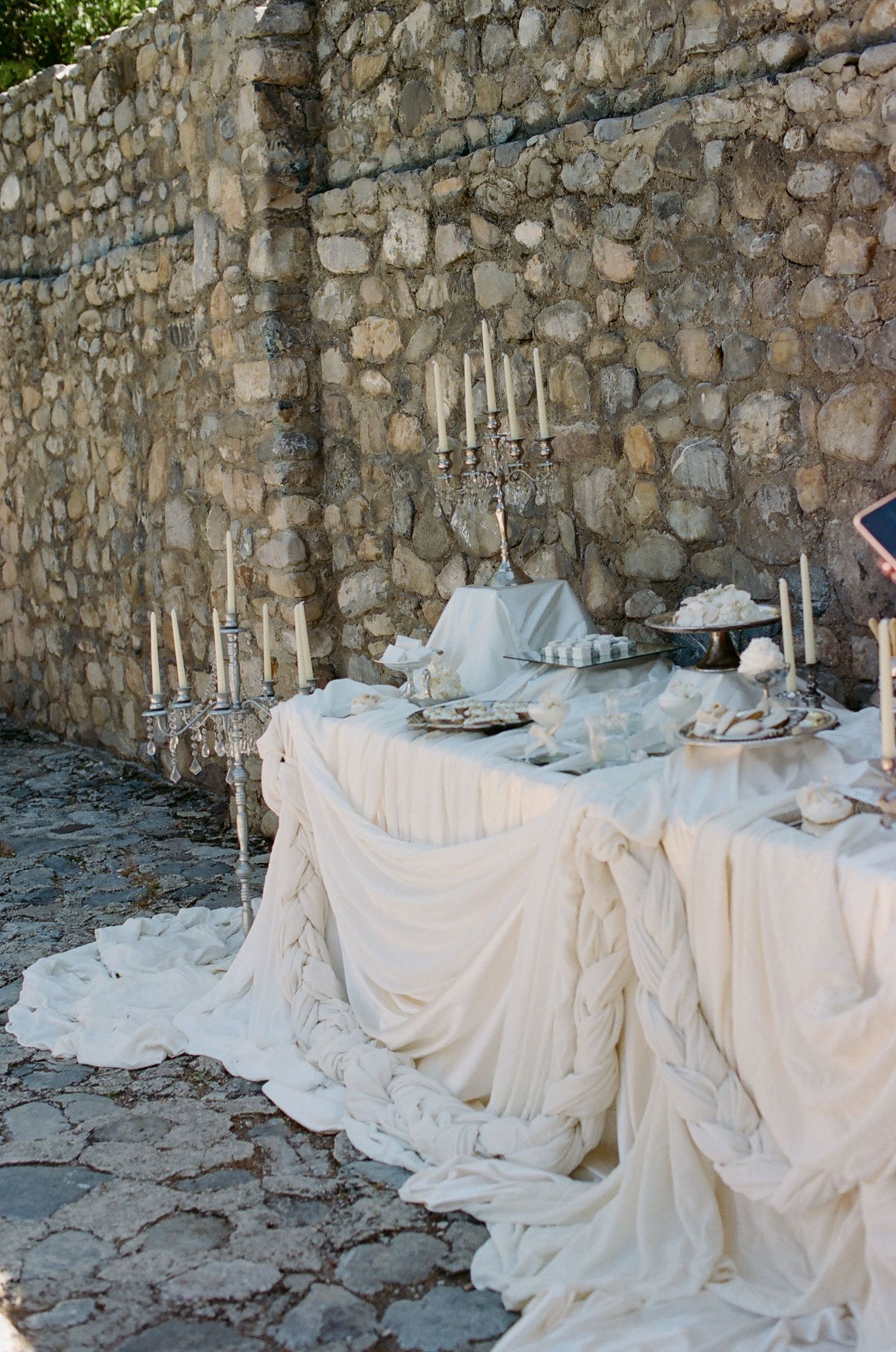 Elegant outdoor wedding table with white draped linen, silver candelabras, and decorative items against a stone wall.