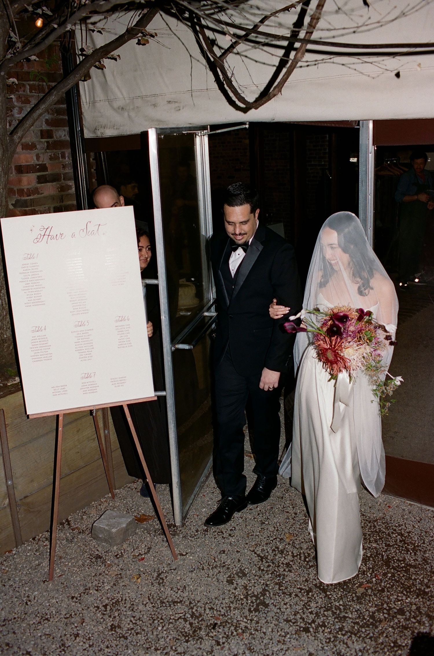 A couple stands next to a seating chart display at their wedding ceremony, with the bride wearing a long veil and white gown.