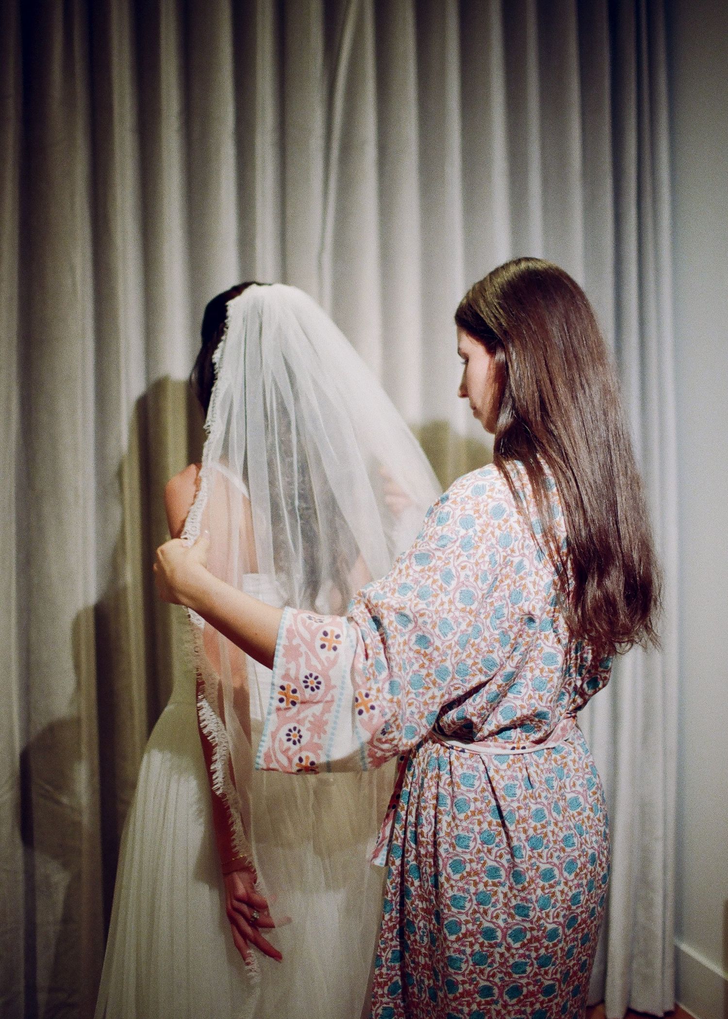 A bridesmaid in a floral robe helps a bride adjust her wedding veil in front of a neutral curtain backdrop.