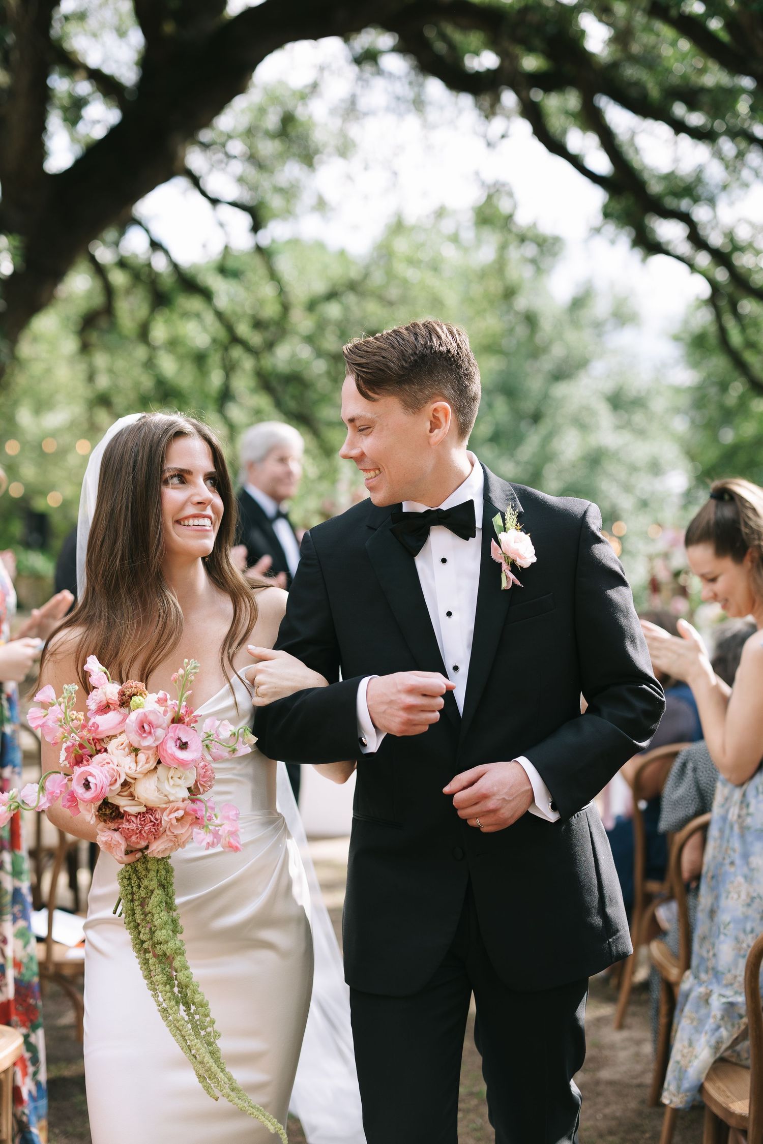 Wedding celebration with pink floral bouquets and formal attire under natural tree canopy.