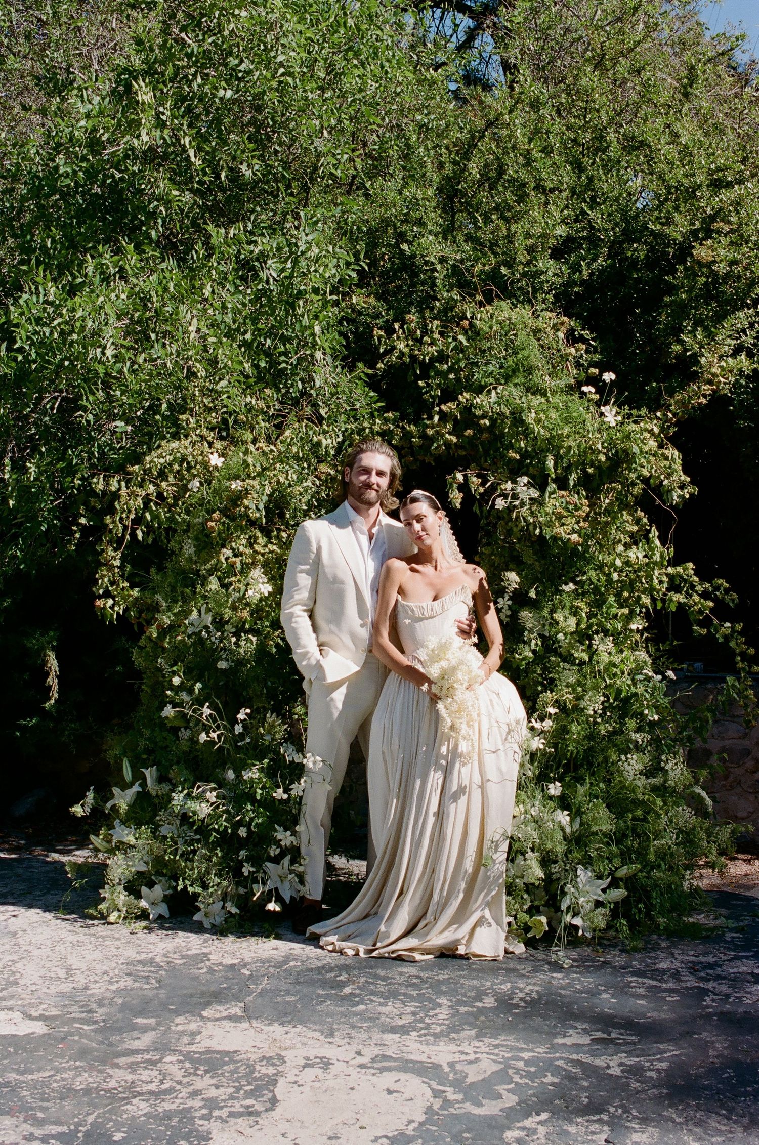 Bride and groom pose together in front of lush green foliage and white floral arch on their wedding day.