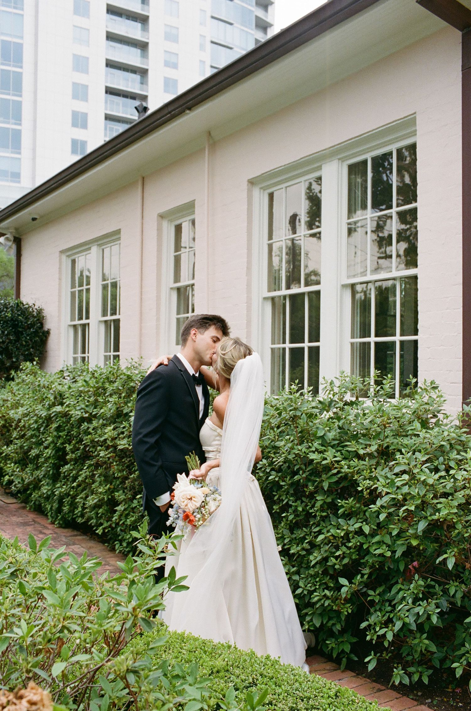 A couple shares a romantic moment outside a white building with lush green landscaping.