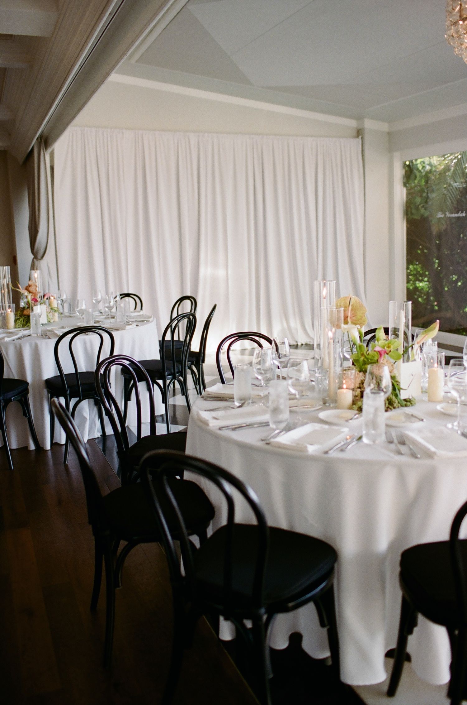Wedding reception venue with white tablecloths and black bentwood chairs against a white curtain backdrop.