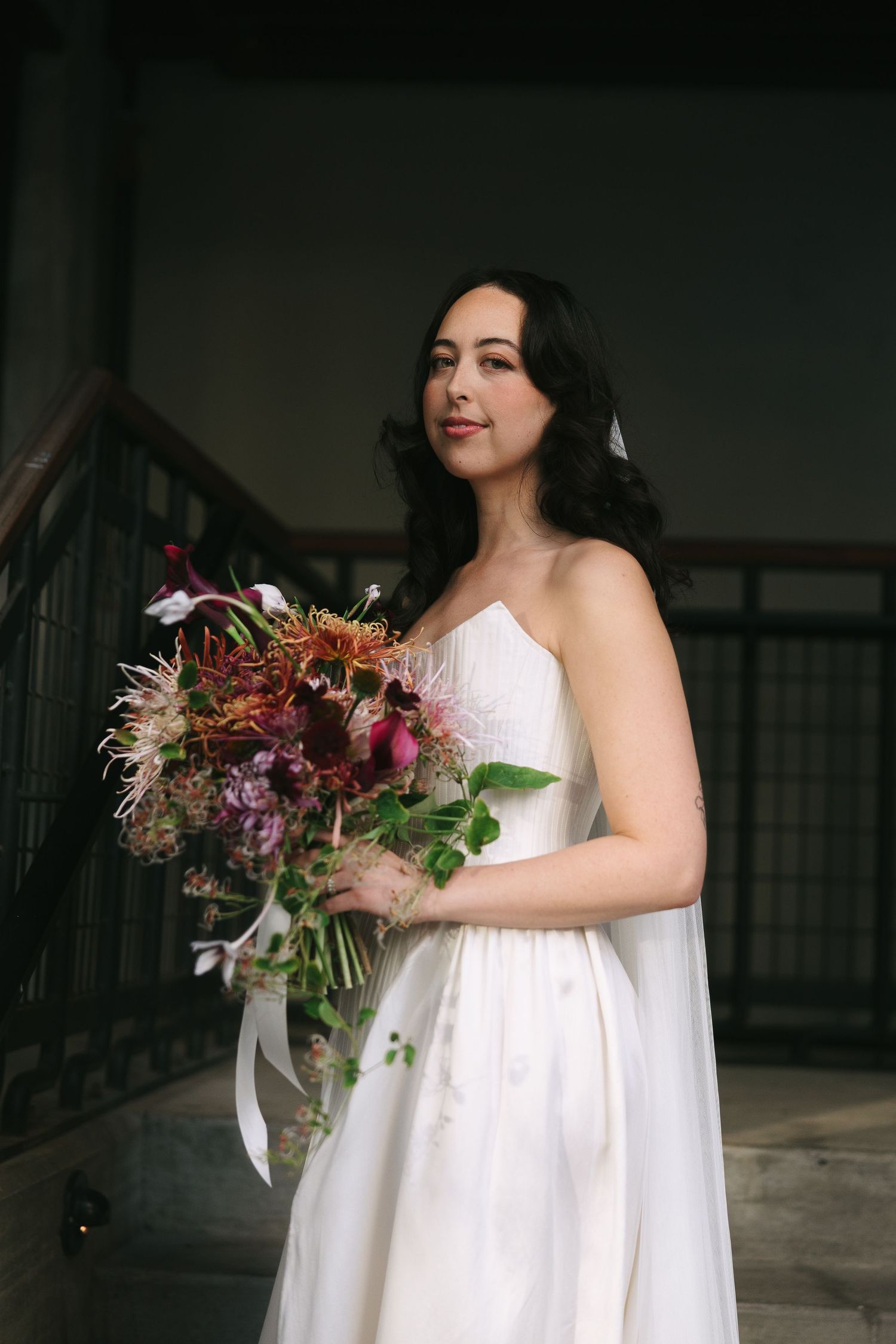 A bride in a strapless white gown poses with a dramatic autumn-colored bouquet on concrete stairs.