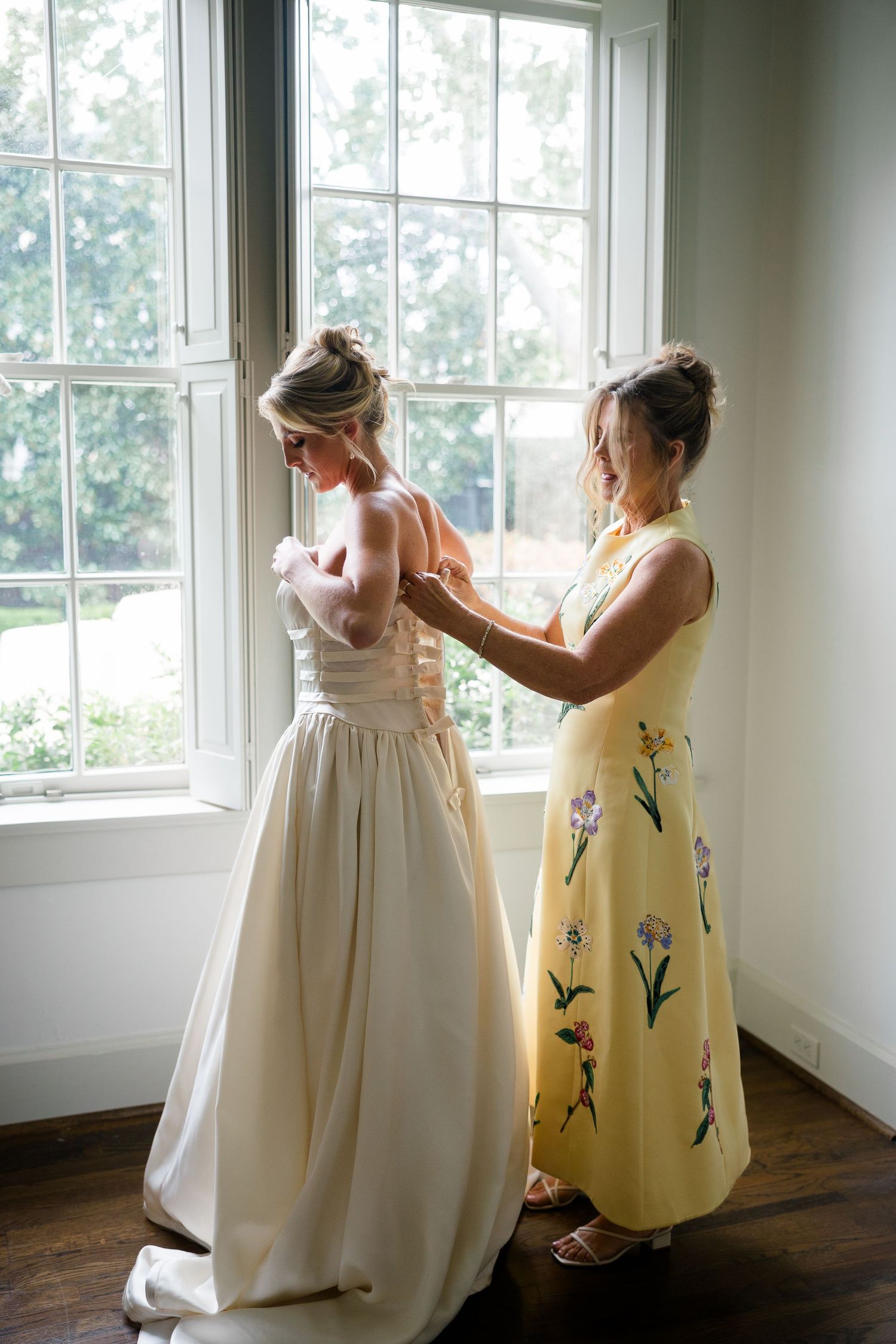 Two people in formal dresses preparing by bright windows.