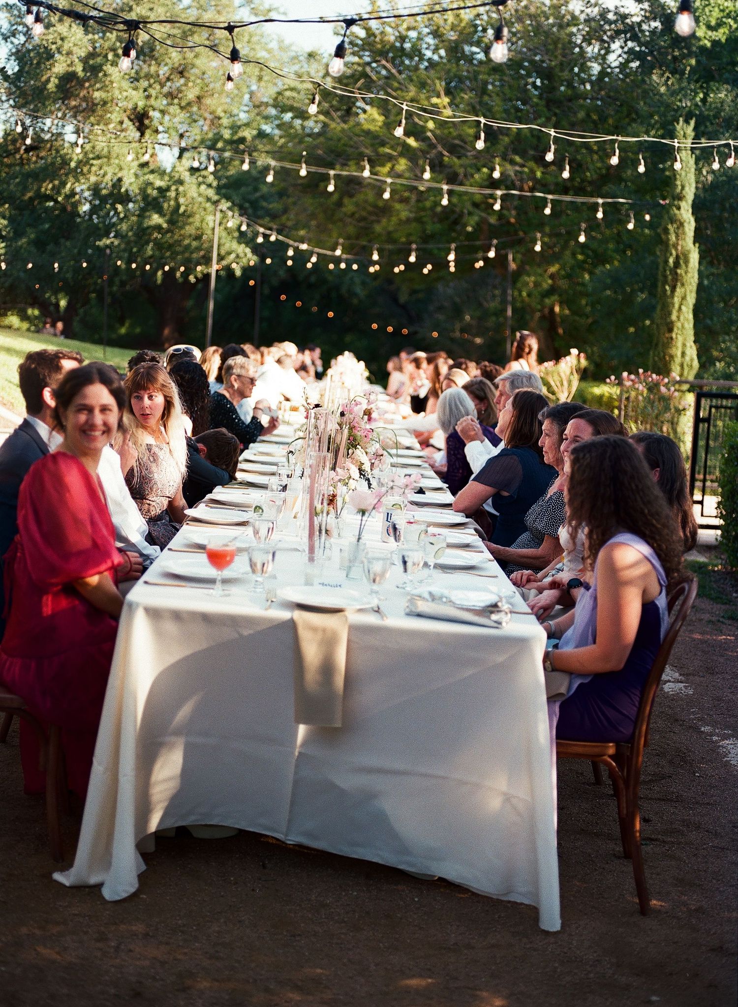 Long tables set for an outdoor wedding reception under string lights at dusk.