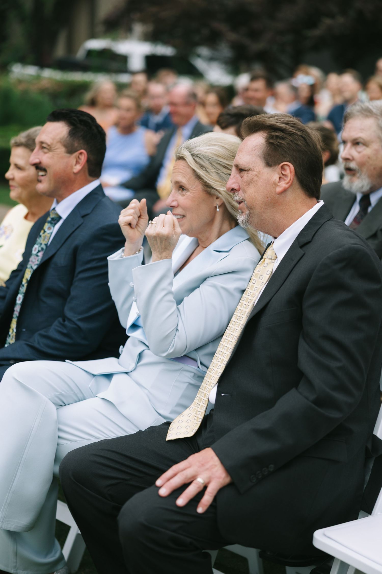 Wedding guests in formal attire seated in rows during an outdoor ceremony.