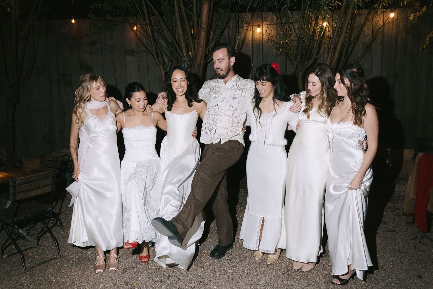 A group poses at a wedding celebration with attendants in white dresses and one person in brown attire.