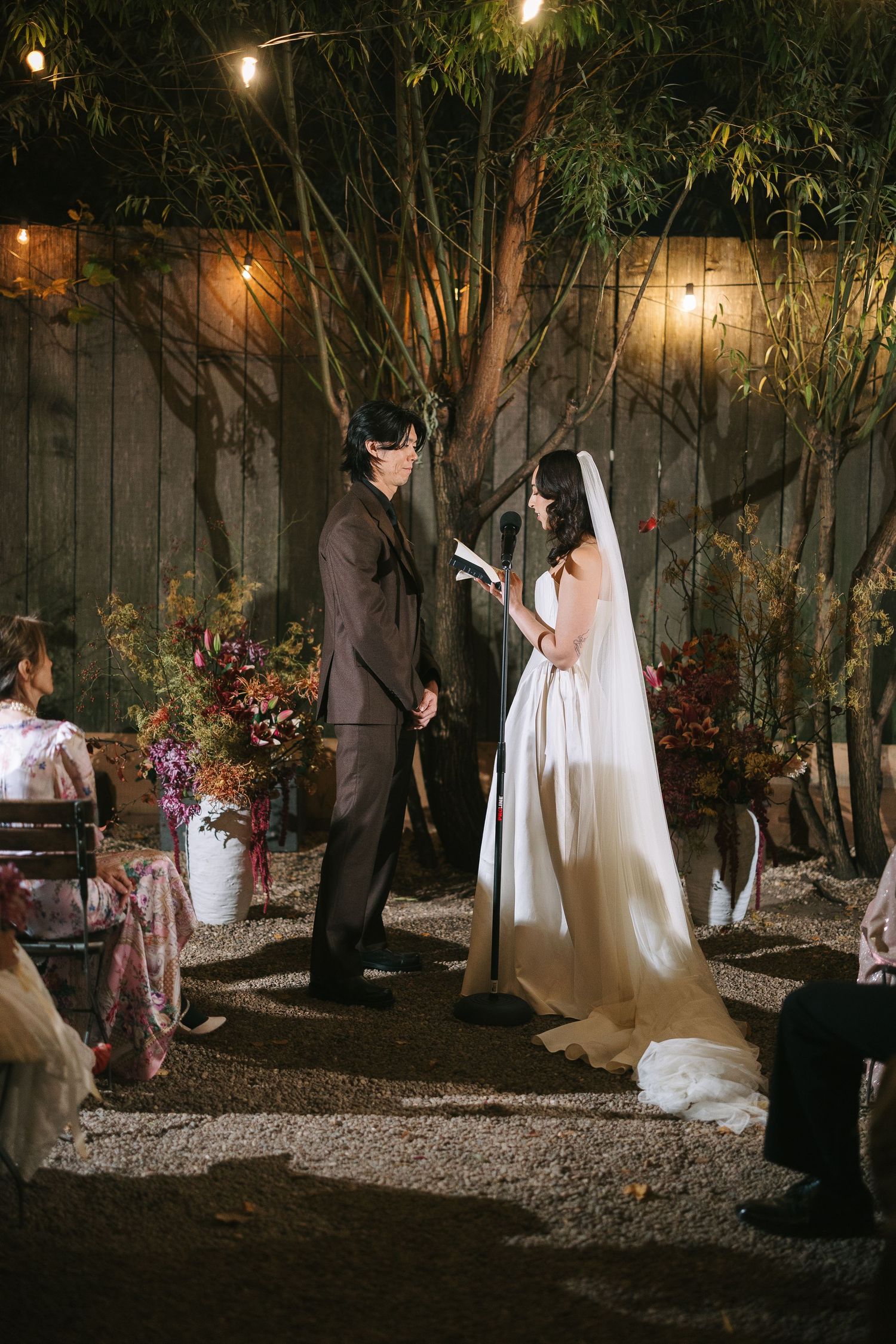 A romantic outdoor wedding ceremony at night with string lights illuminating a garden setting.