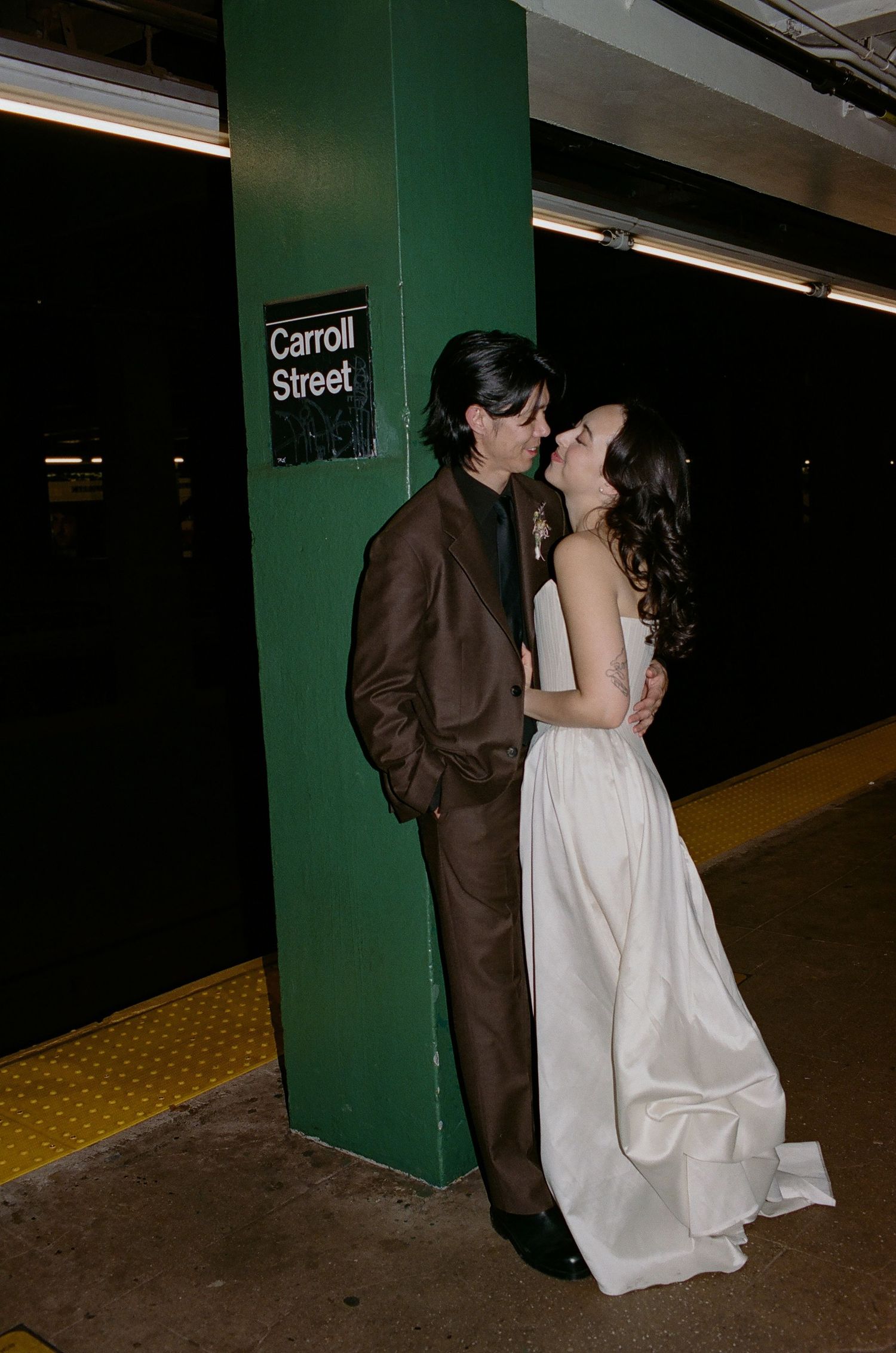 A romantic couple in formal attire share an intimate moment at a subway station platform near a green pillar.
