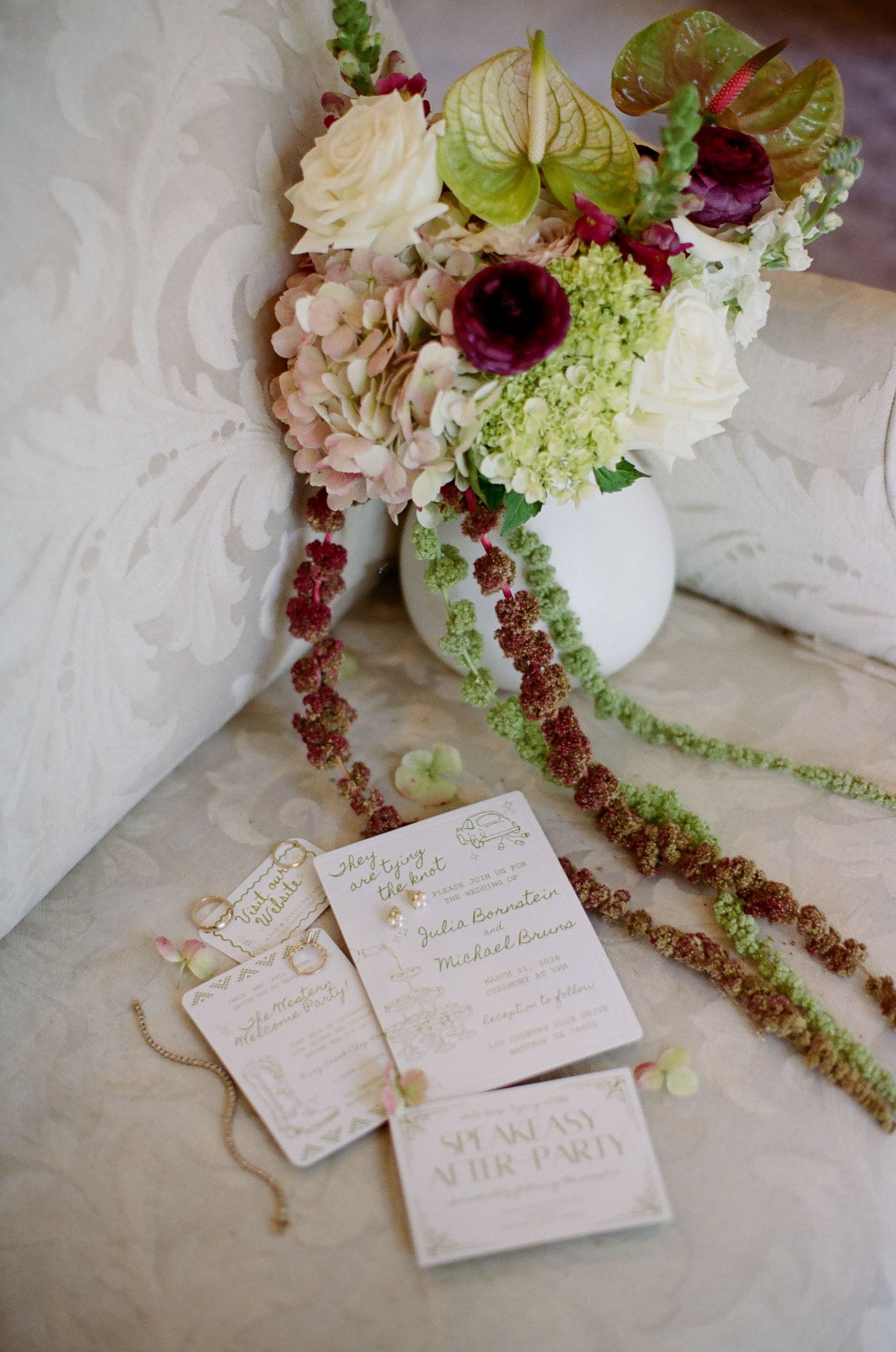 Elegant wedding invitation suite with floral arrangement featuring burgundy, cream, and green blooms in white vase.