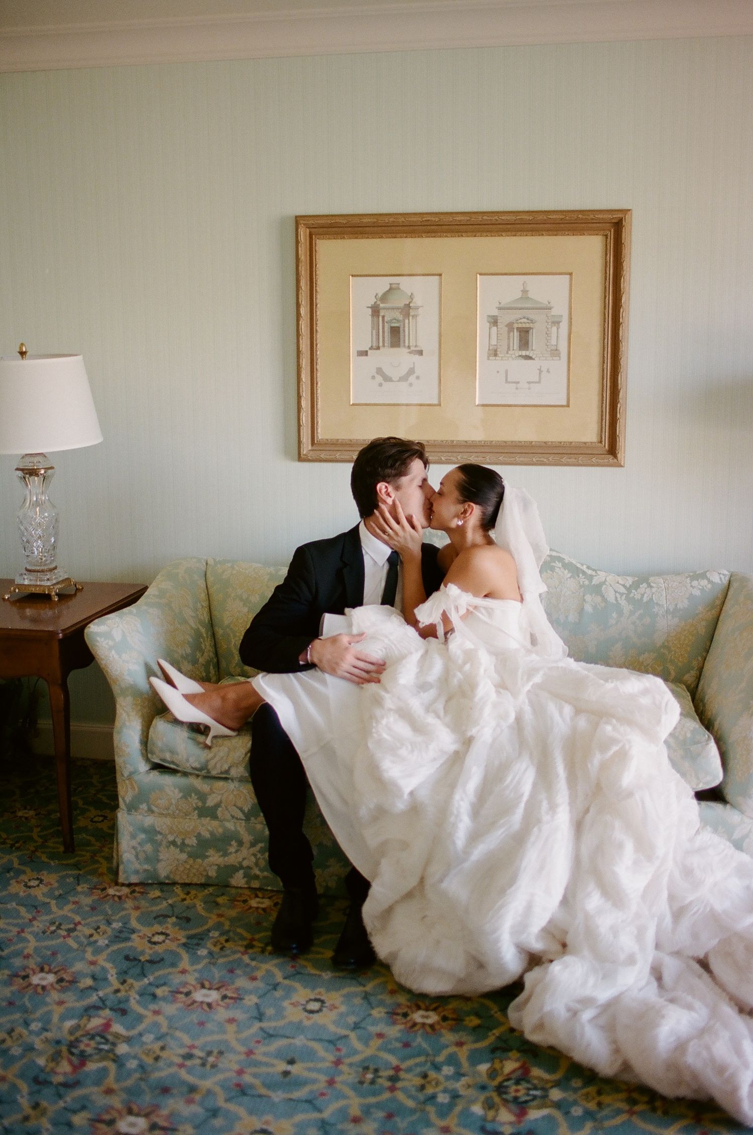 Bride and groom share a romantic kiss on a floral sofa, bride in a voluminous white gown with veil.