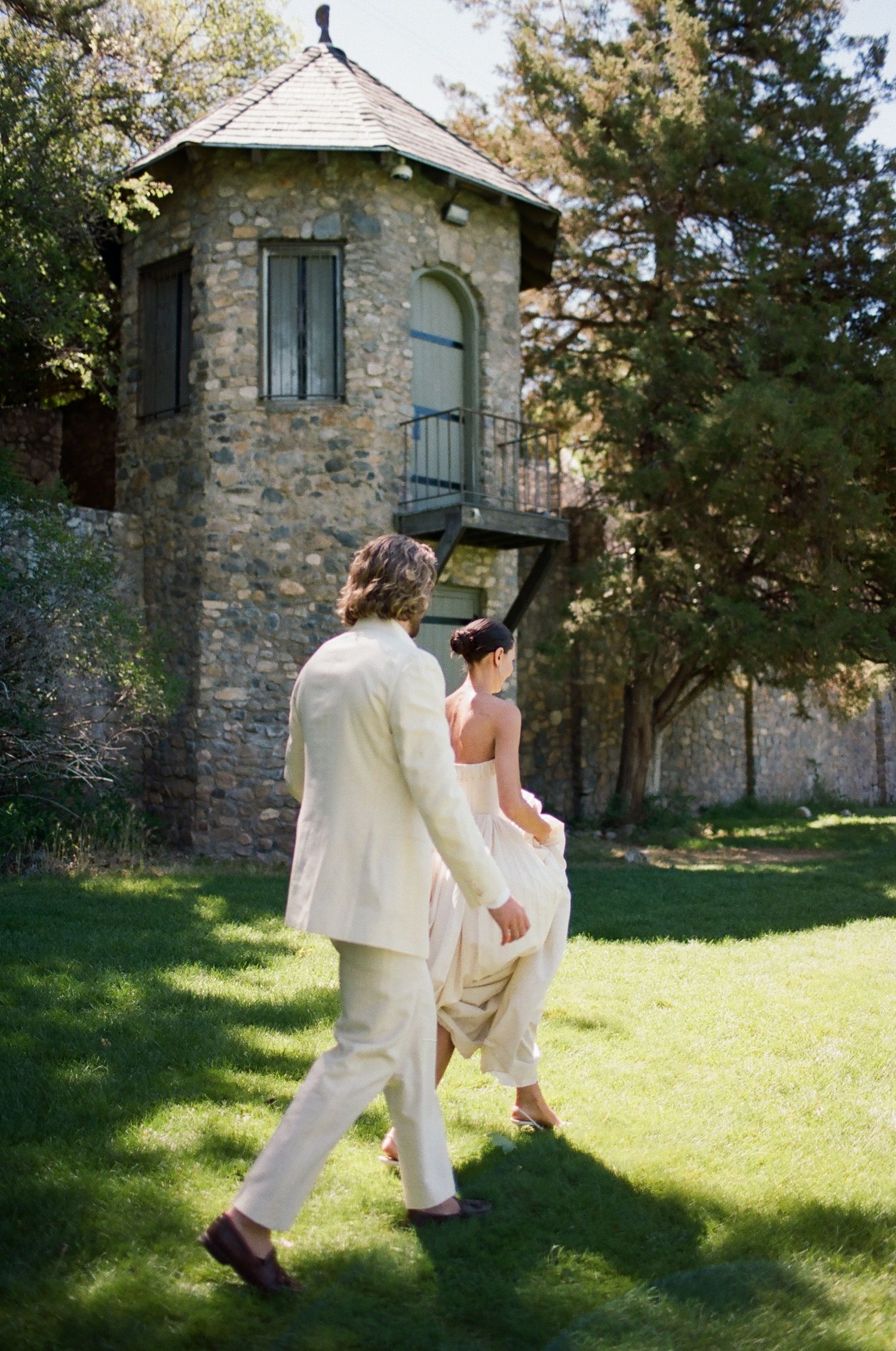 Bride and groom walking hand in hand across lush green lawn toward a rustic stone tower on a sunny wedding day.