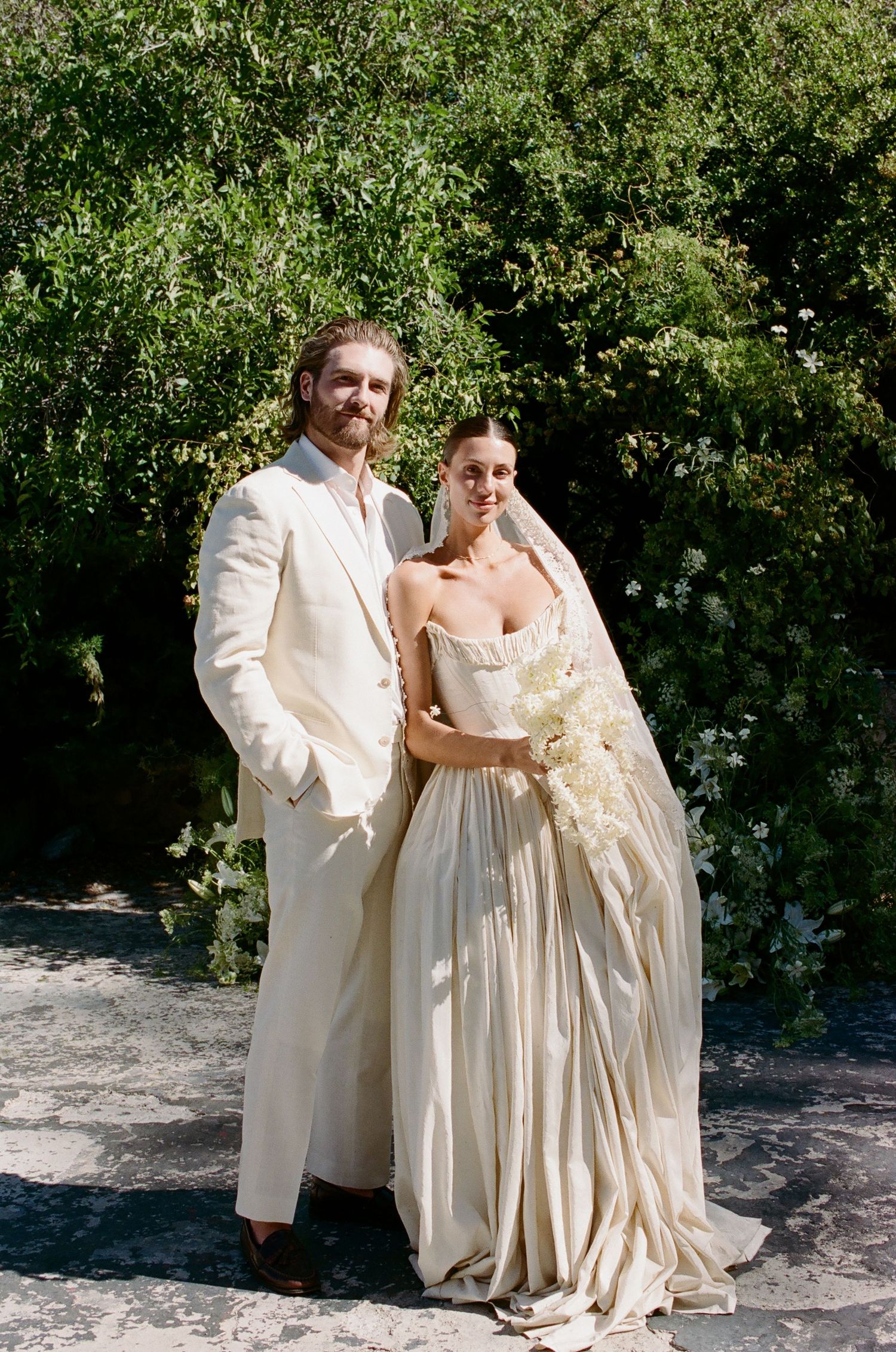 A couple poses outdoors in elegant cream wedding attire, bride holding white florals with lush greenery backdrop.