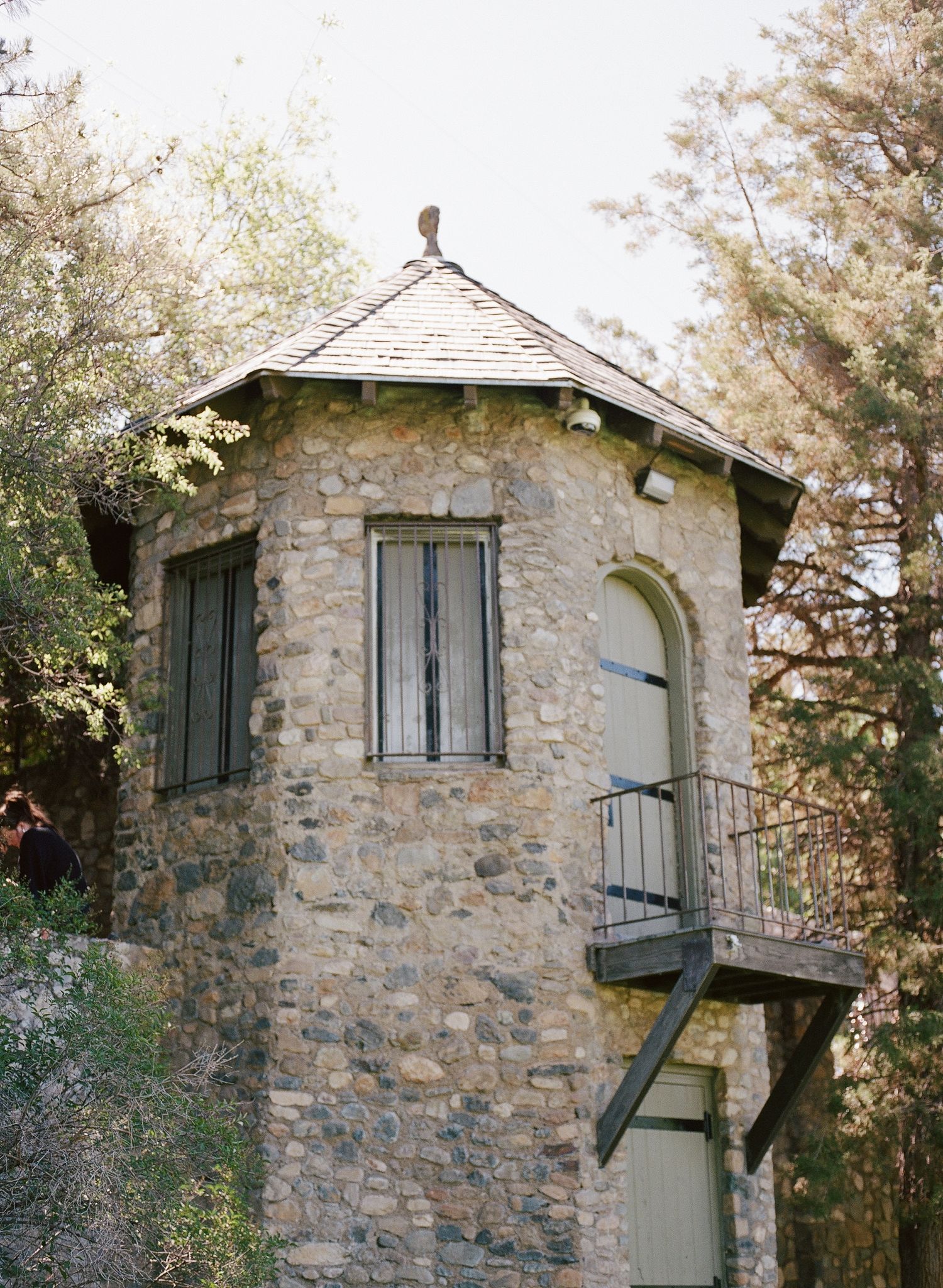 Historic stone tower with arched window, slate roof, and metal balcony surrounded by tall trees.