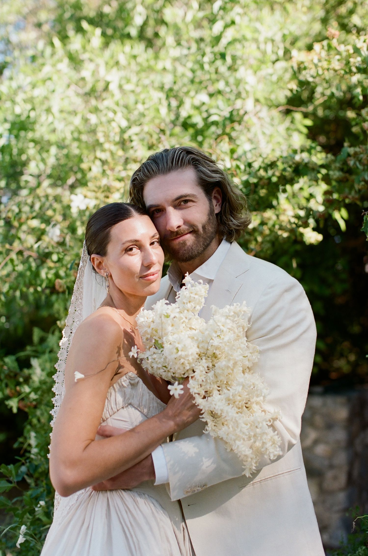 Bride and groom embrace outdoors holding white floral bouquet, wearing elegant wedding attire with lush green trees behind.