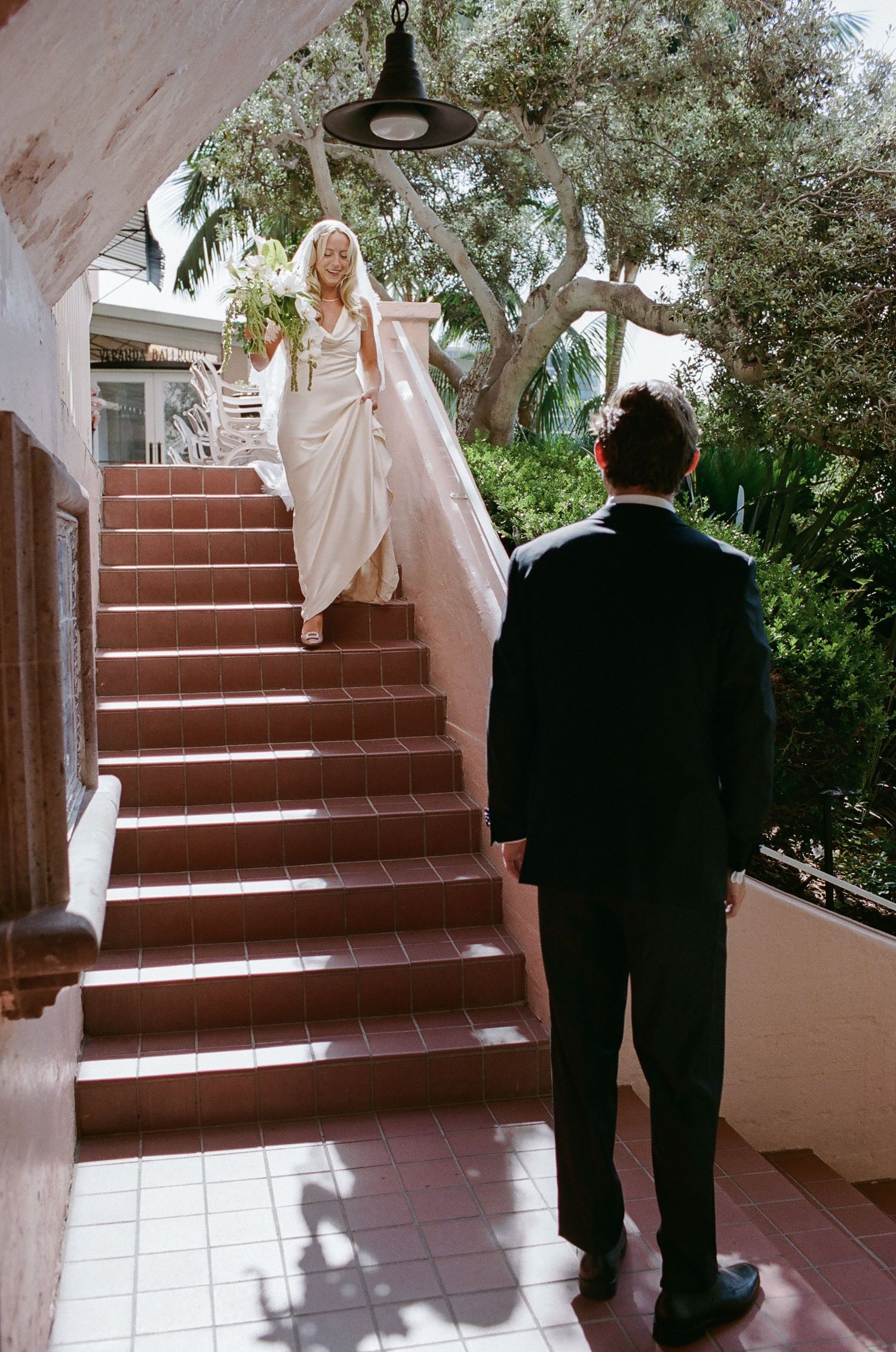 Bride in white gown descends red-tiled stairs toward groom in black suit at outdoor wedding venue.