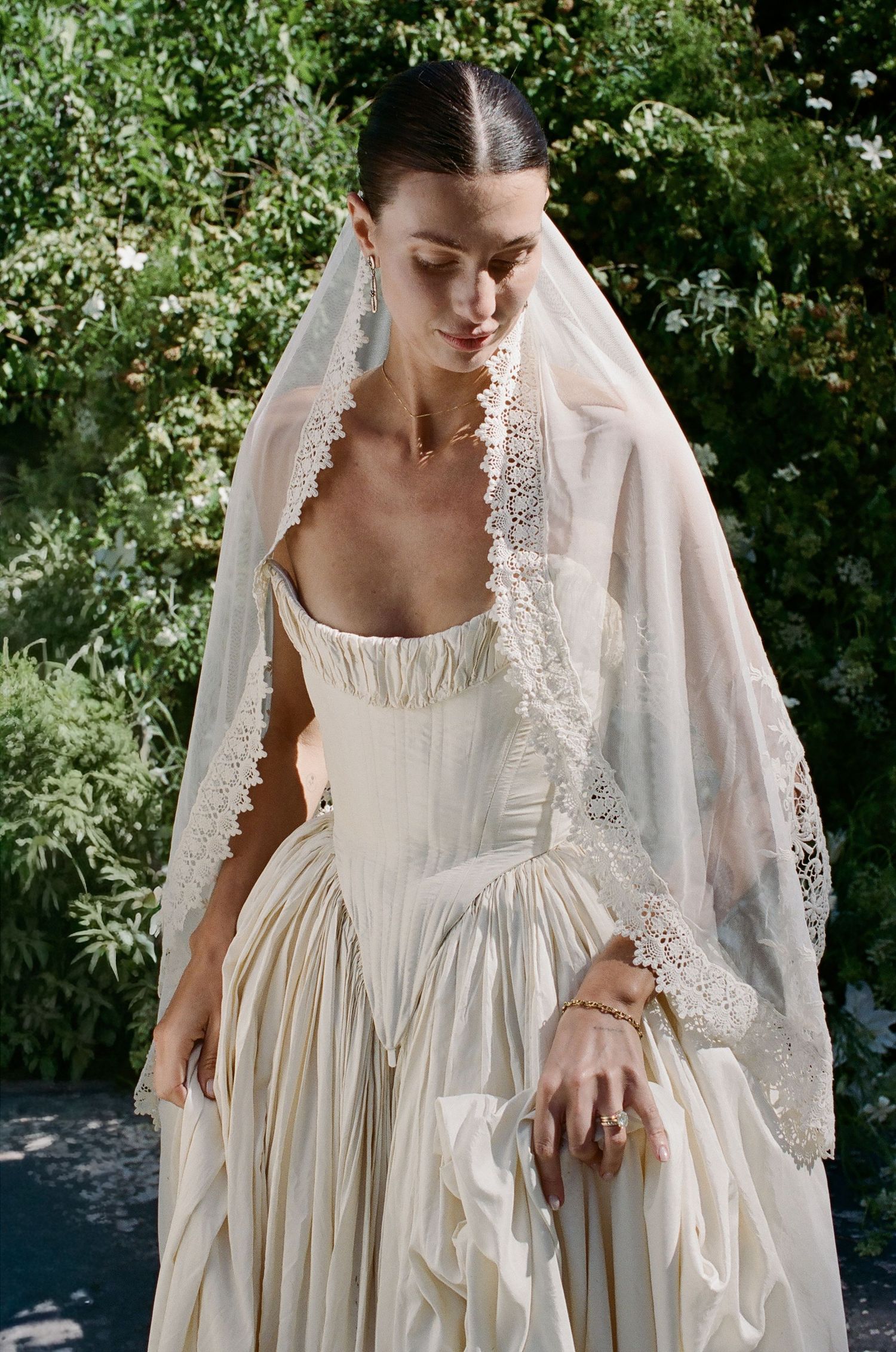 Bride wearing ivory corset gown with lace-trimmed veil, standing outdoors among lush green foliage.