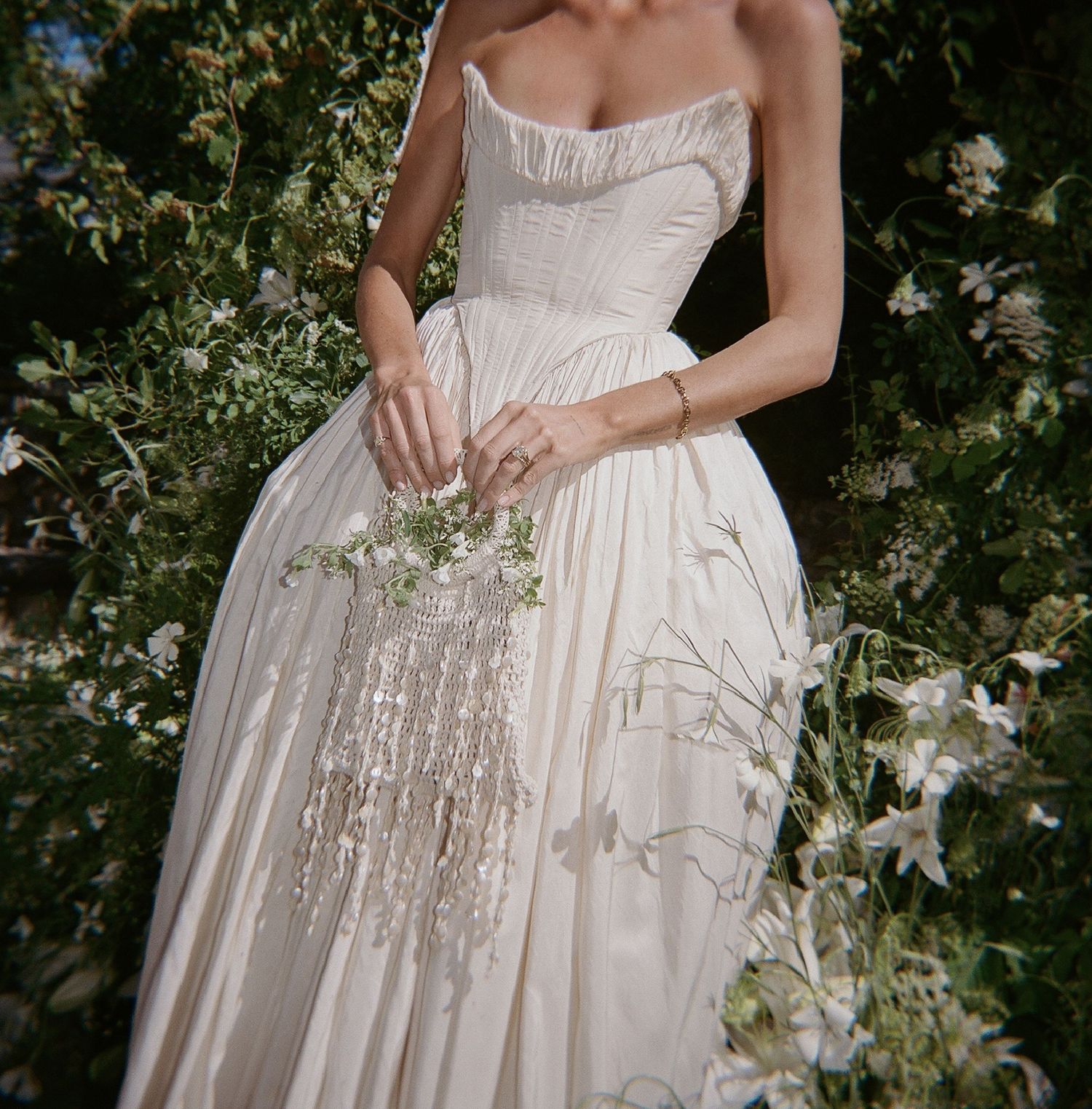 Bride in strapless ivory ball gown holding delicate wildflower bouquet surrounded by lush white garden blooms.