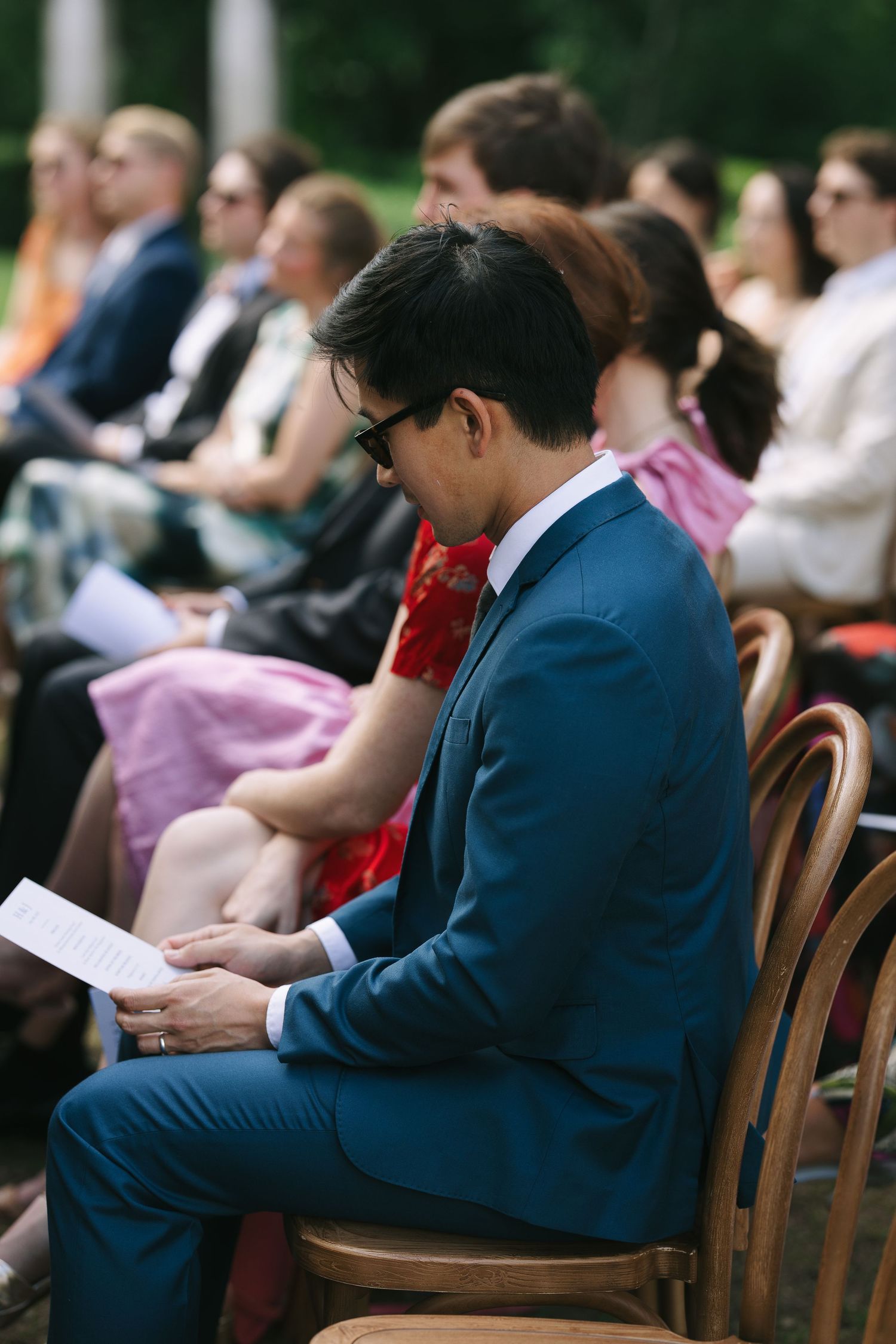 Wedding guests reading ceremony programs while seated in wooden chairs.