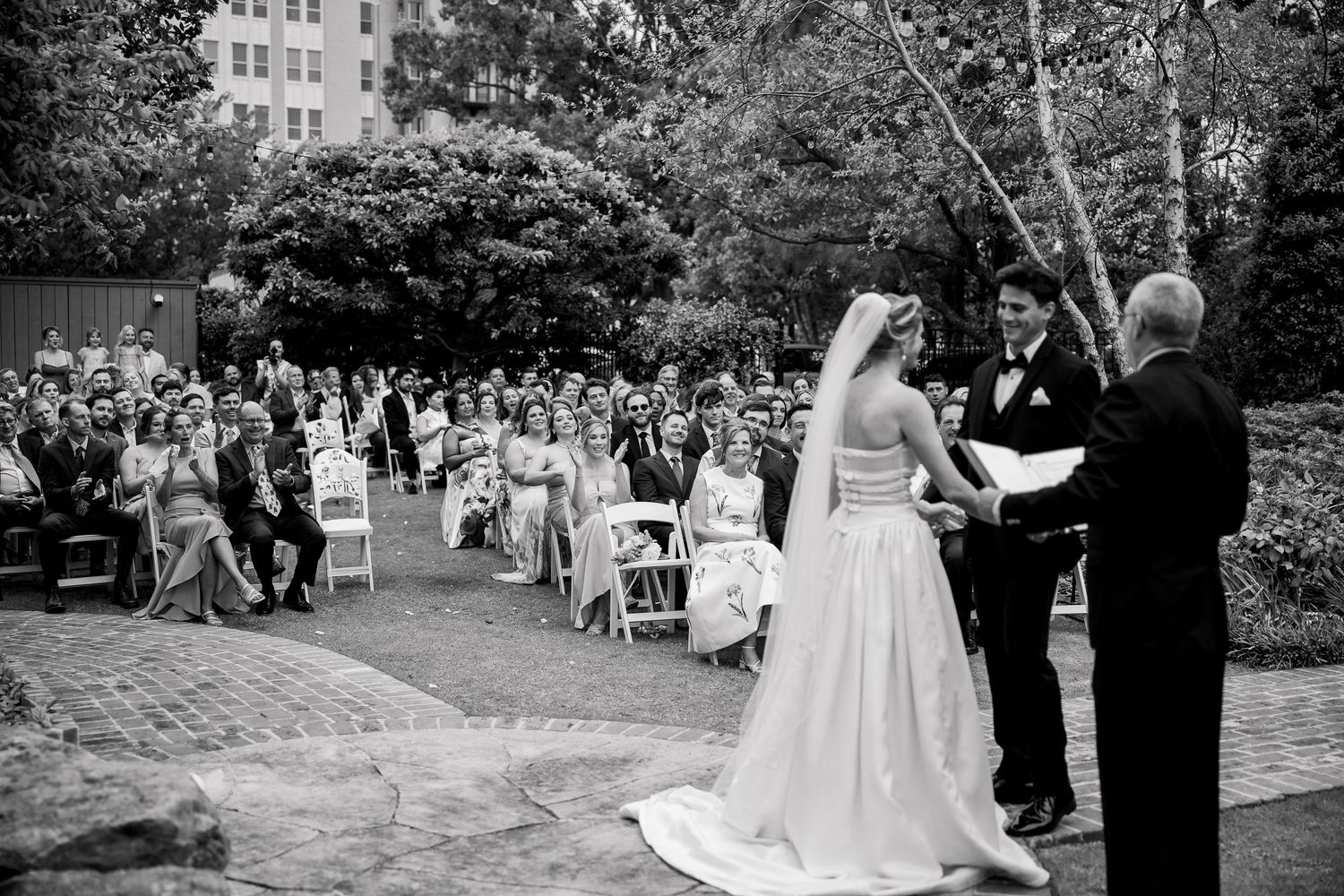 Wedding ceremony in progress on a brick pathway with guests seated outdoors.