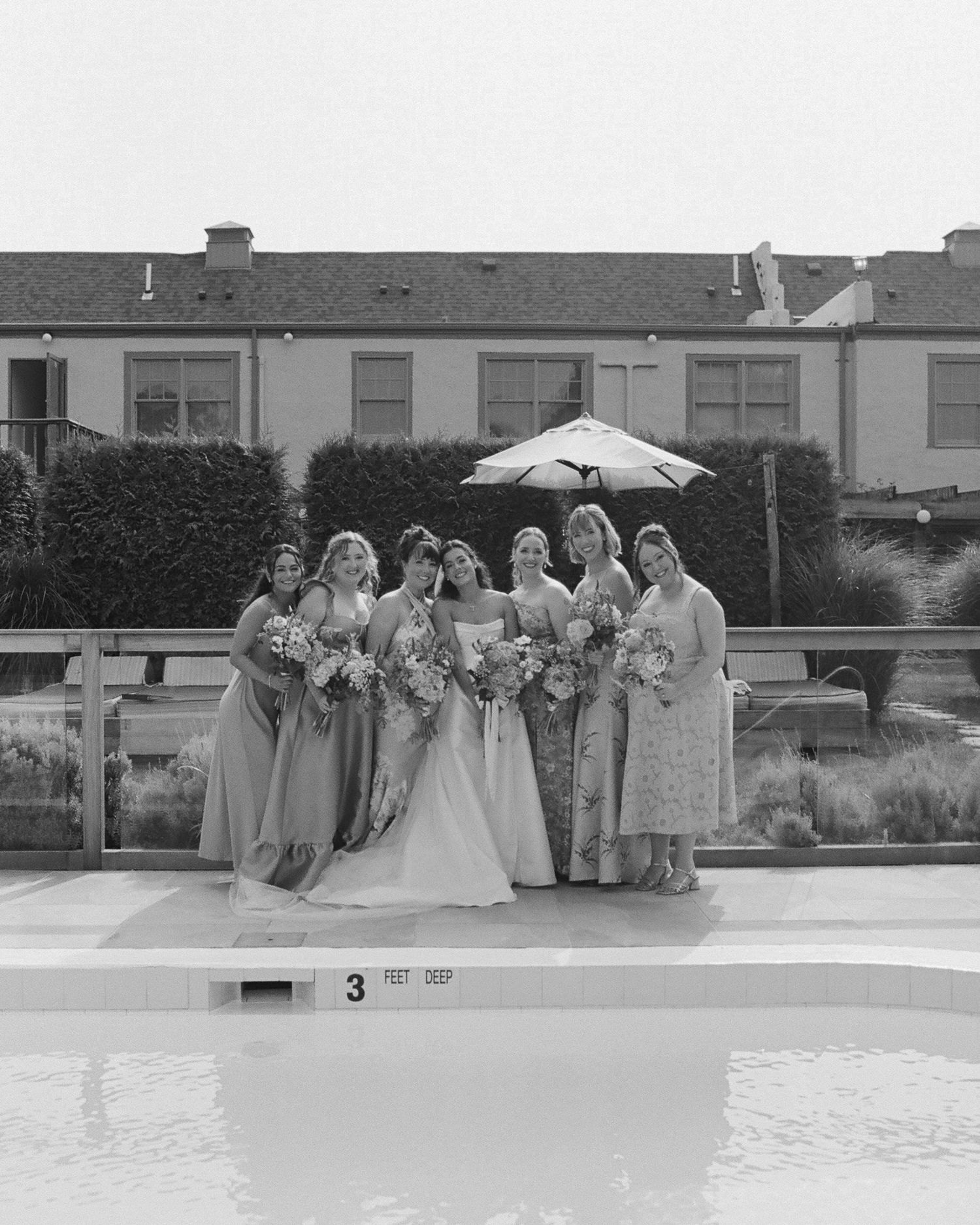 Wedding party poses by poolside in formal attire under cloudy skies.