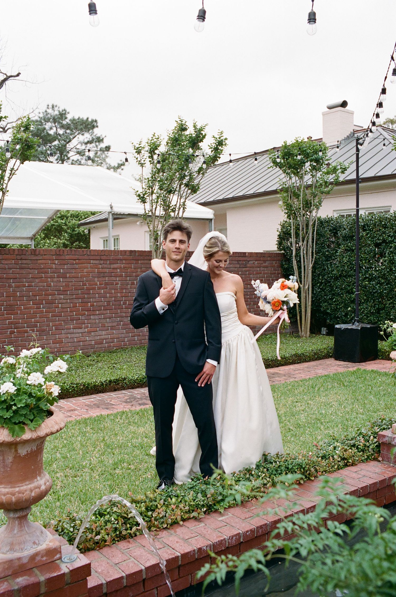 Wedding couple laughs together in an outdoor garden with brick walls and greenery.