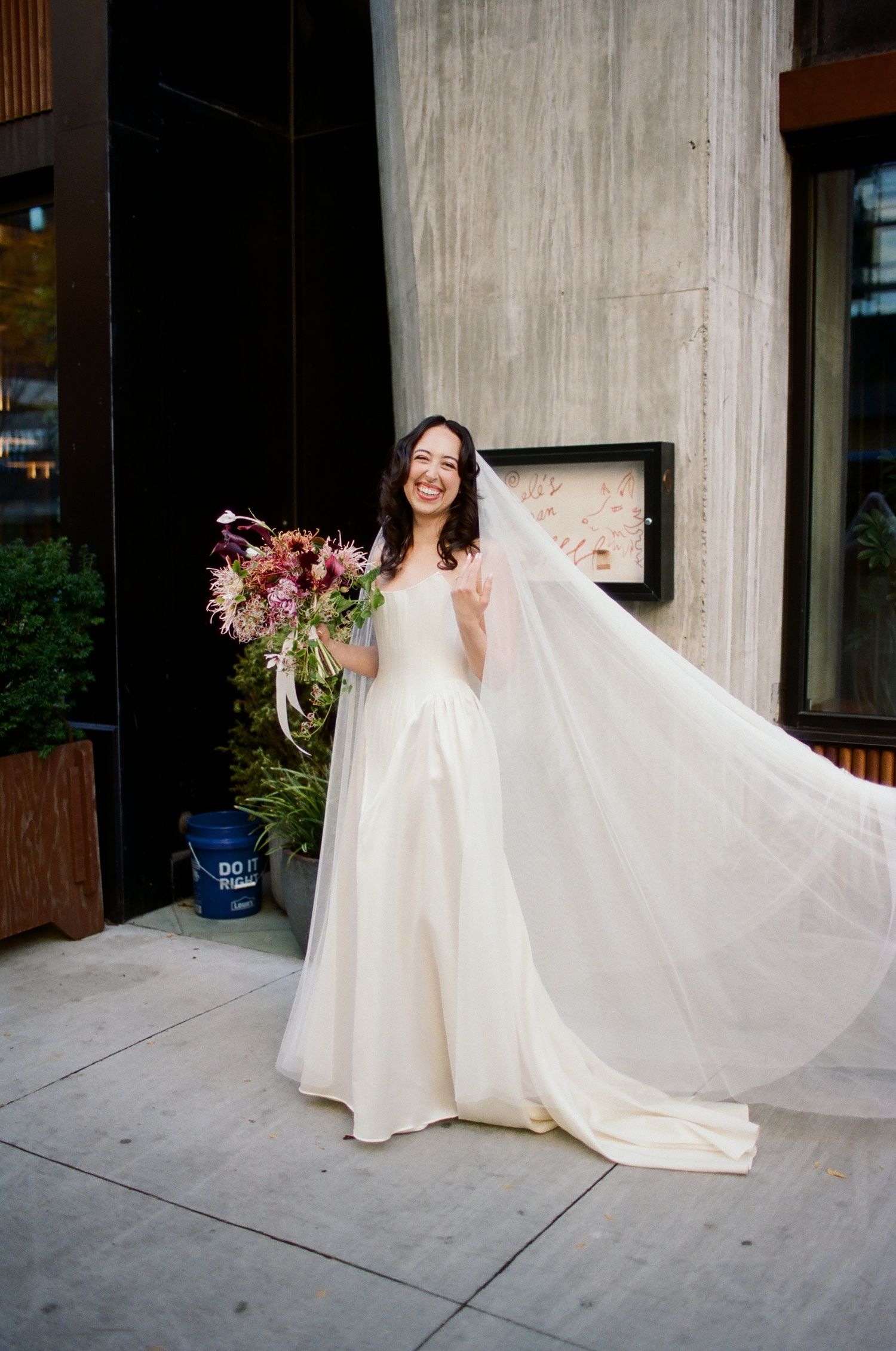 Smiling bride in white gown and flowing veil holds colorful bouquet outdoors, making peace sign.