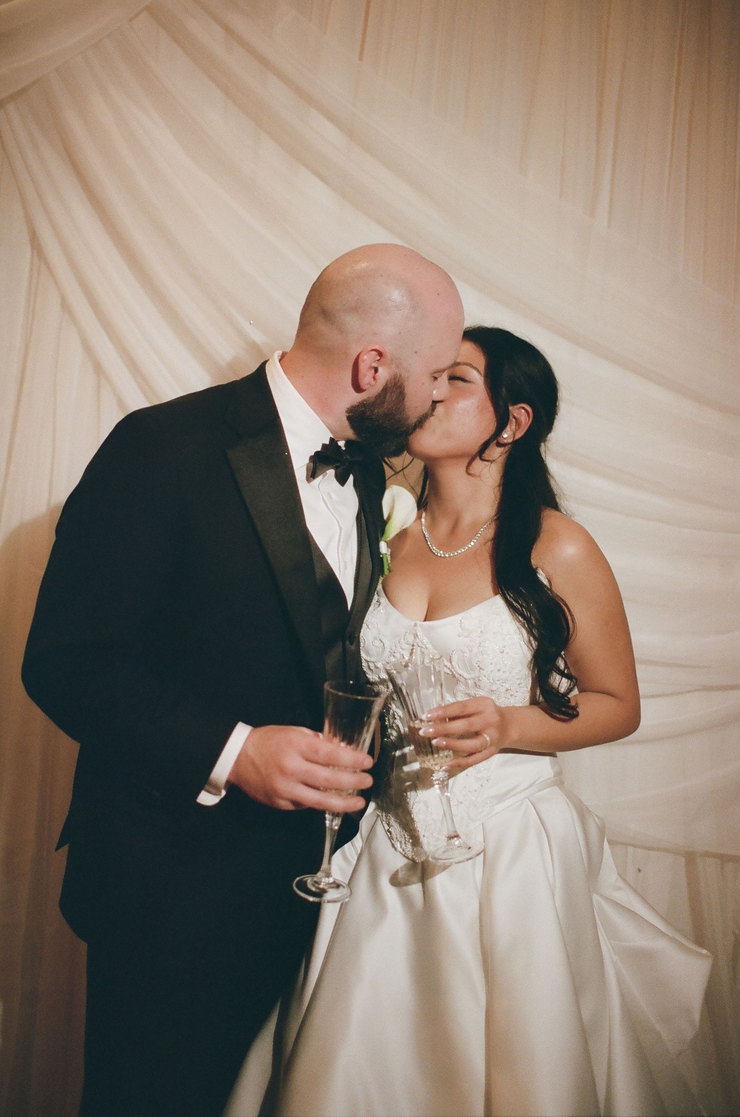 A romantic wedding moment as newlyweds share a kiss while holding champagne glasses against a draped fabric backdrop.