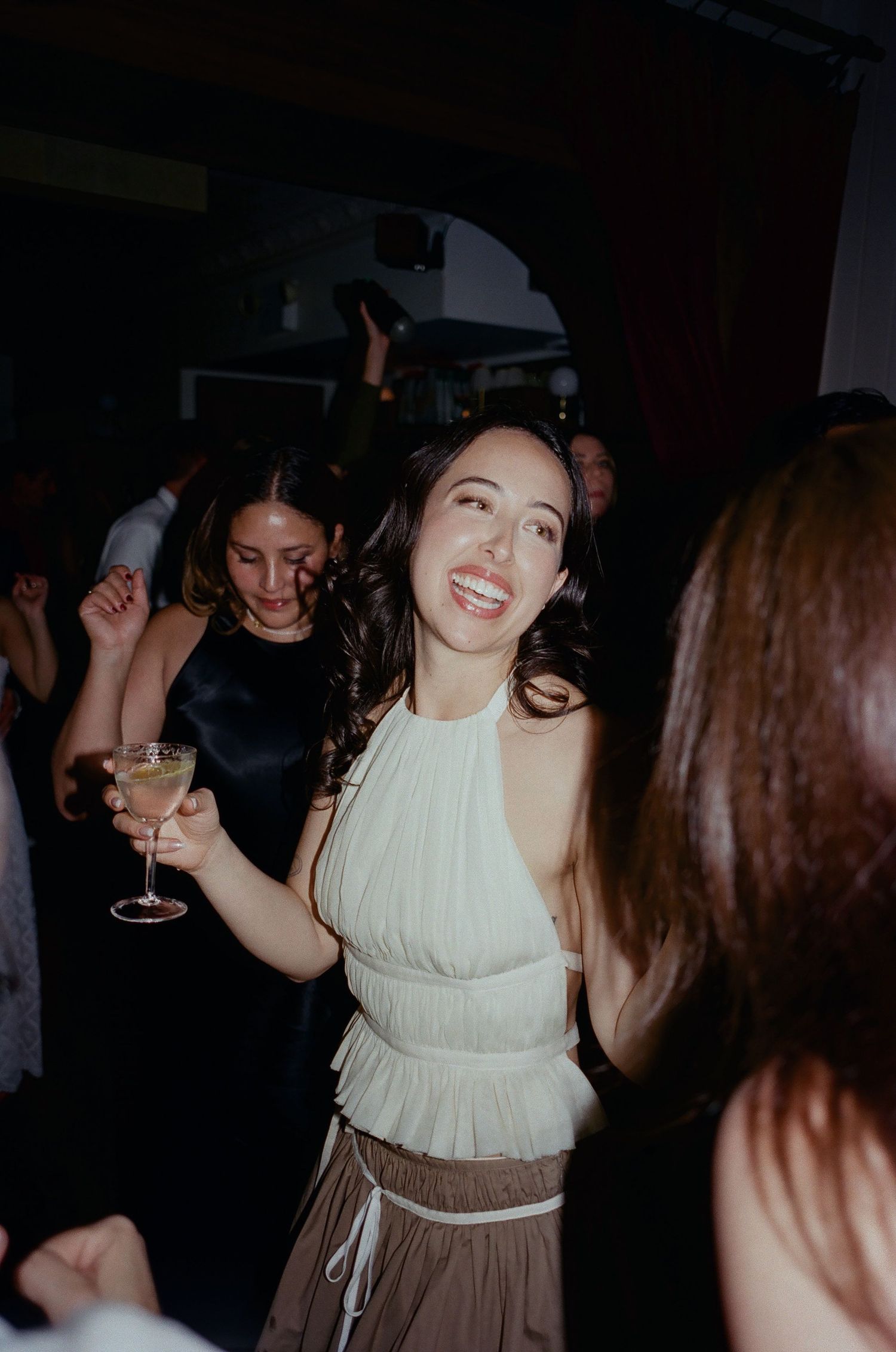People dancing and celebrating at a nighttime party with someone wearing a white halter top and brown skirt holding a drink.