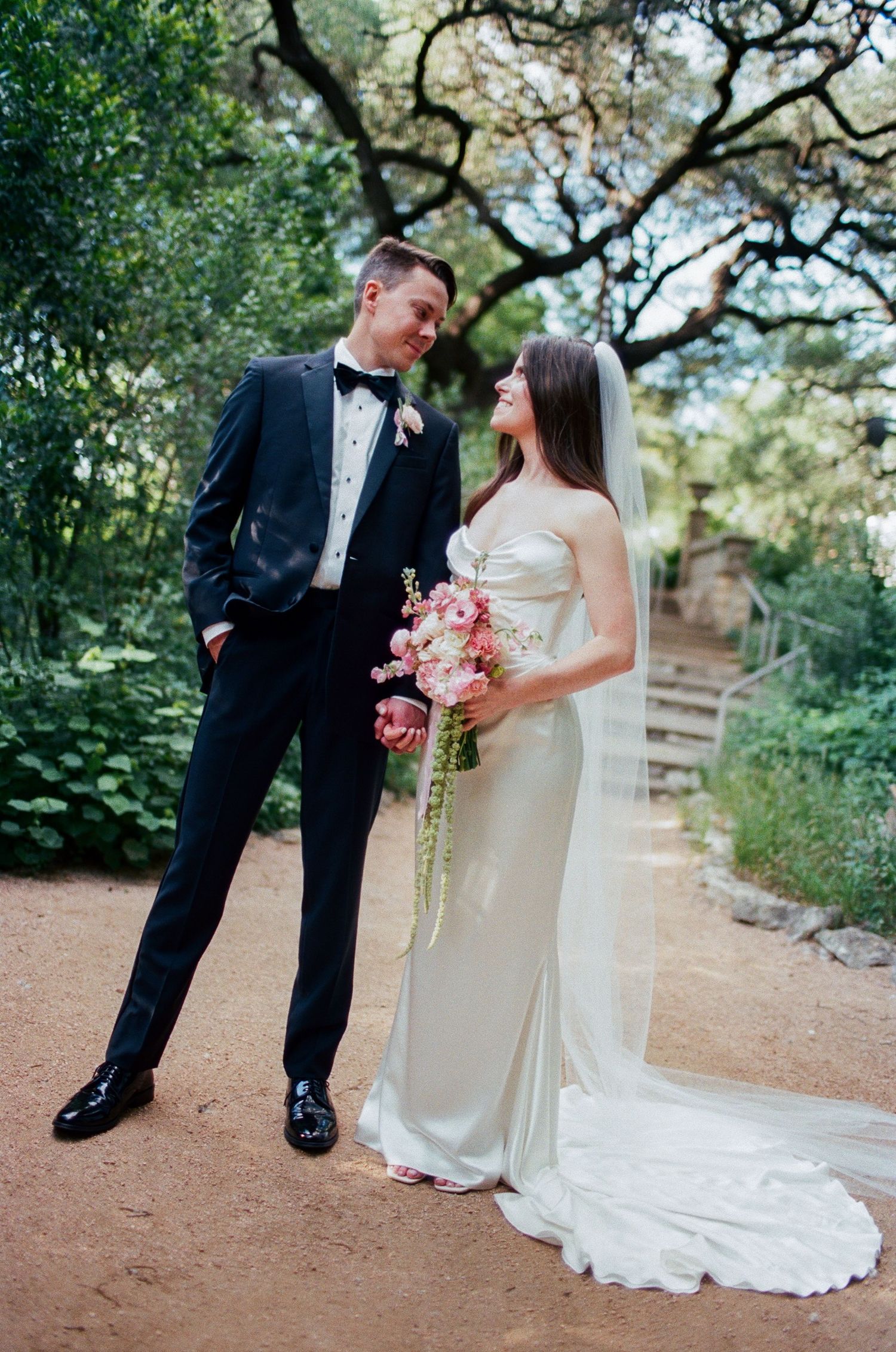 Wedding couple poses on dirt path under overhanging tree branches.
