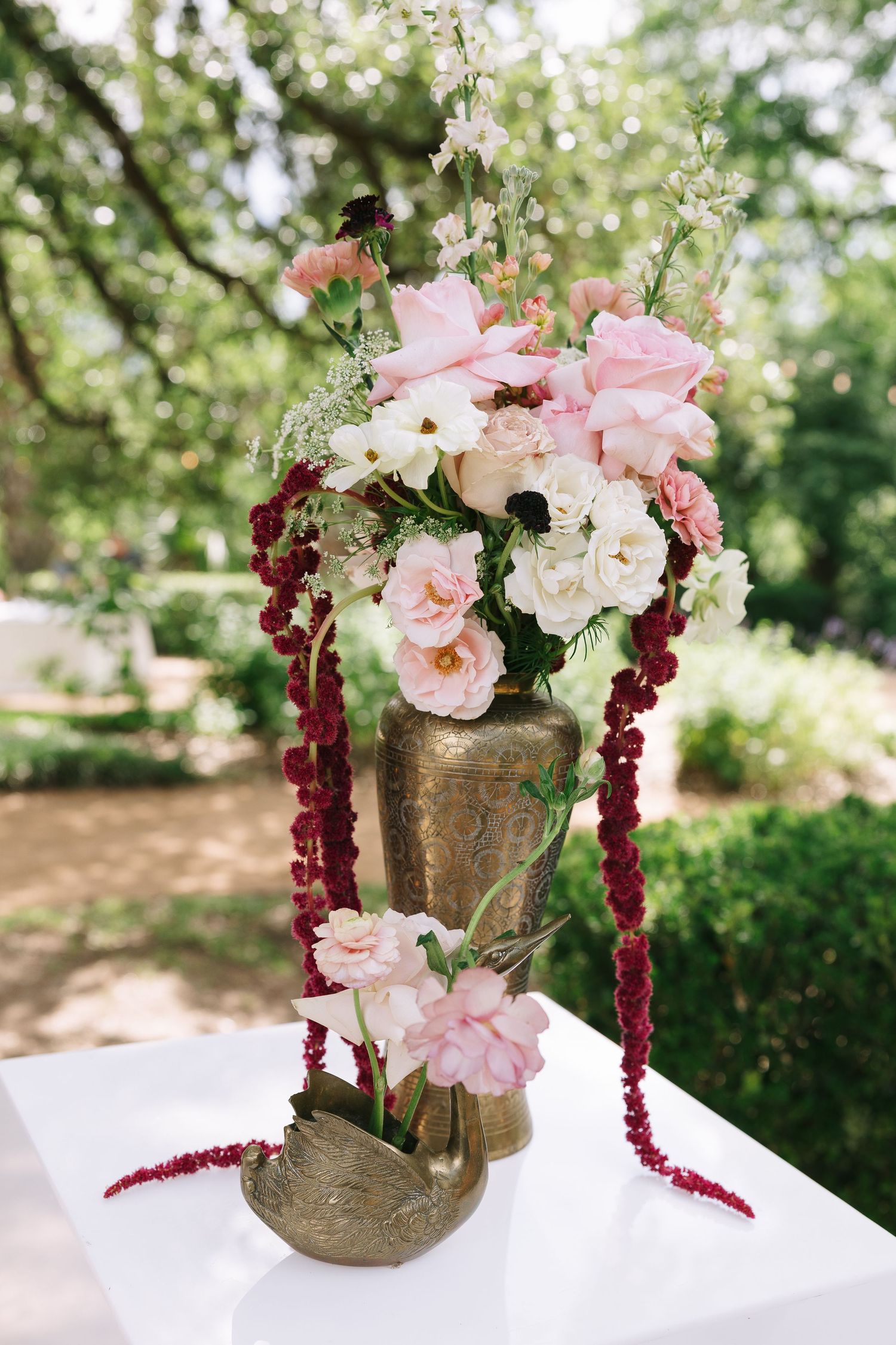 Pink and burgundy floral centerpiece in a gold vase sits atop a white pedestal outdoors.
