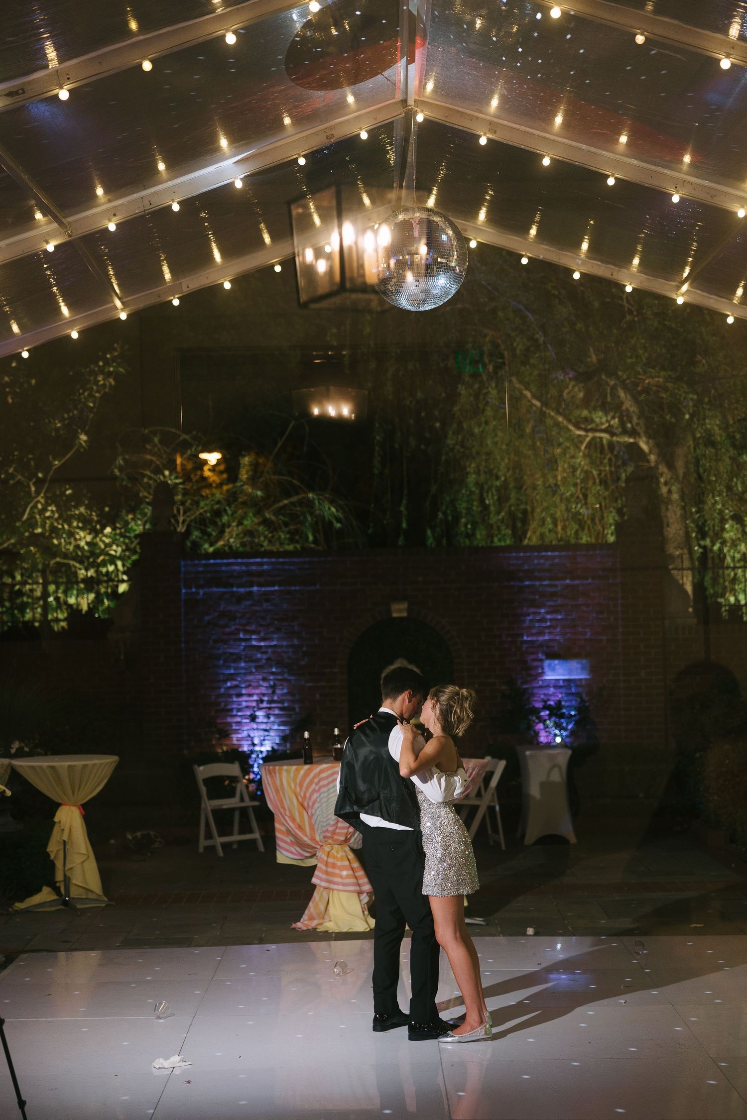 A couple shares a romantic dance under a sparkling disco ball and string lights at an elegant indoor venue.
