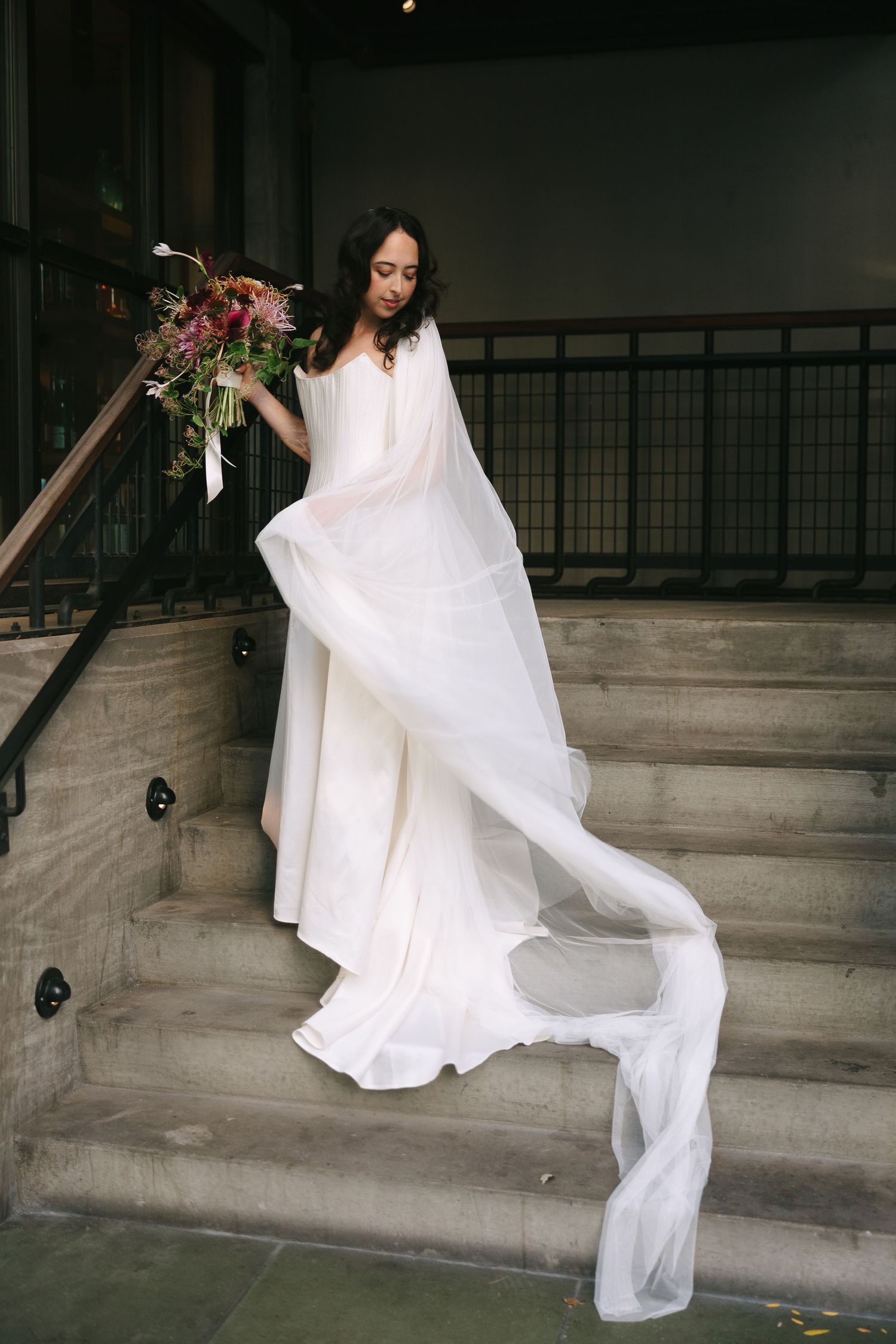 A flowing white wedding dress and veil captured in movement on concrete stairs.