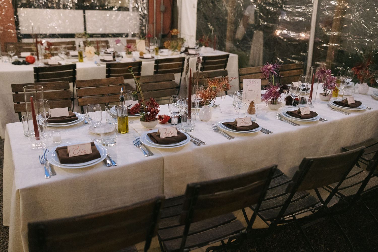 Long banquet table with white linens and place settings arranged outdoors with wooden chairs.