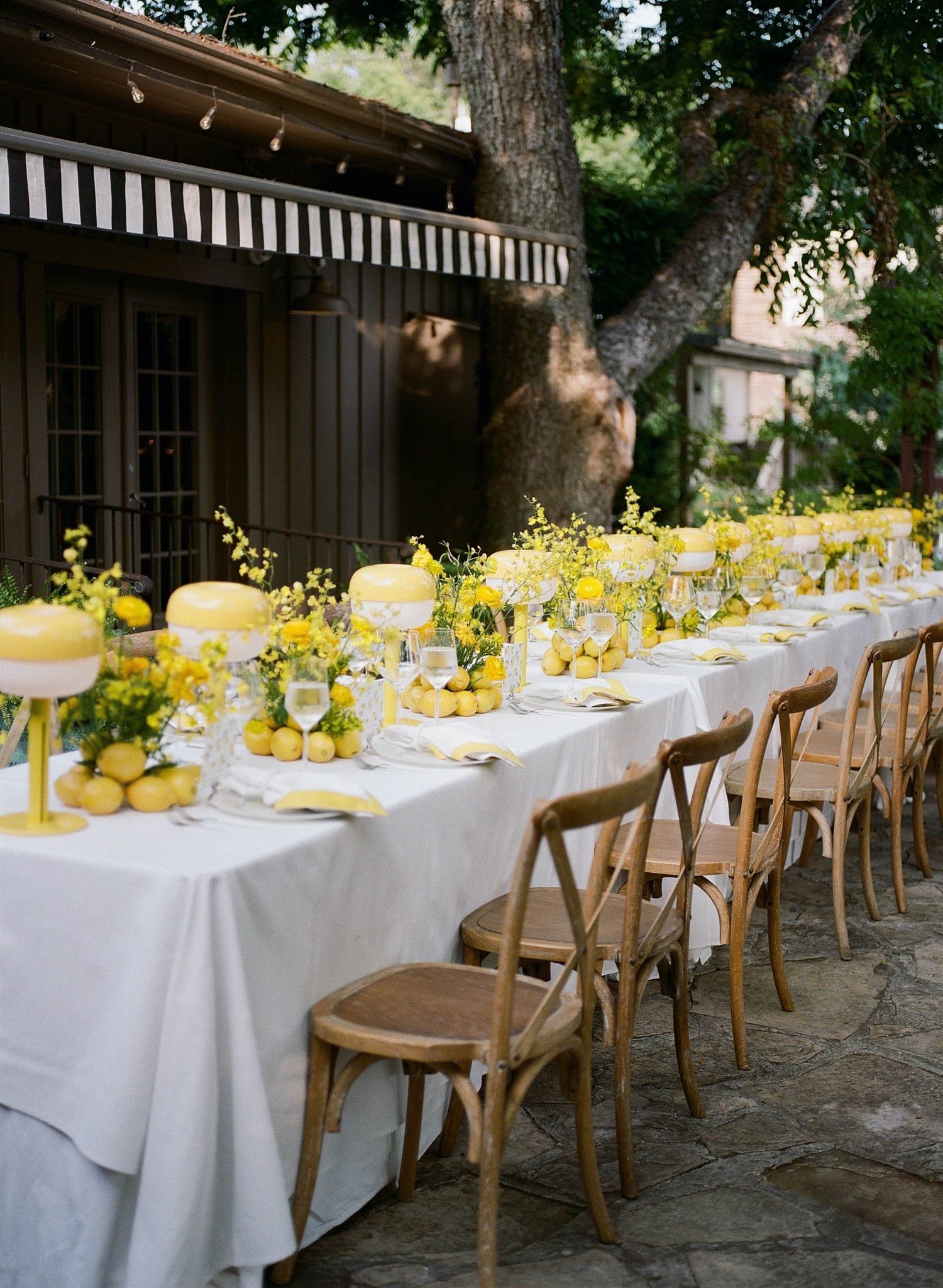 Long white dining tables decorated with yellow centerpieces and wooden chairs set up outdoors under trees.