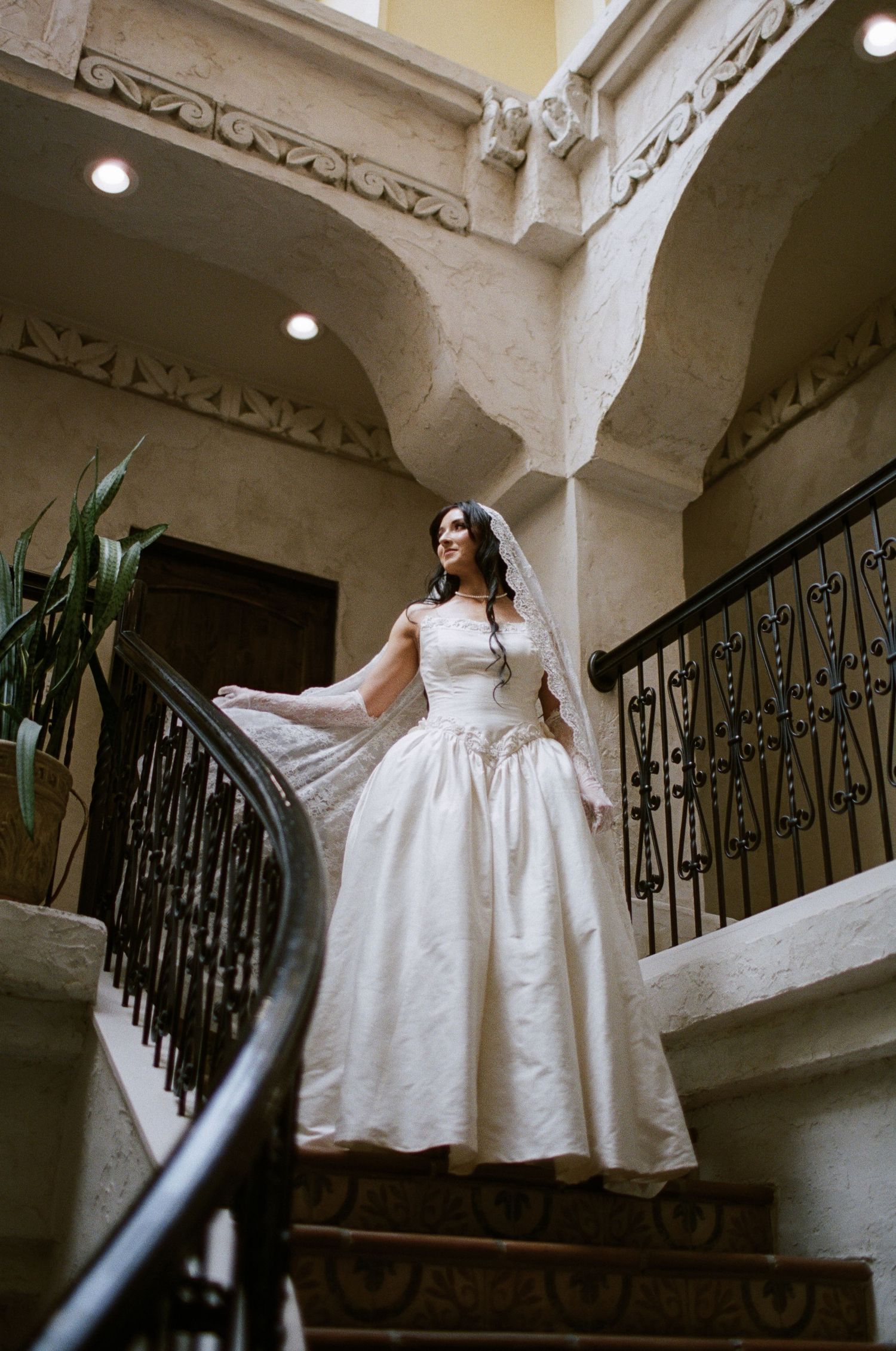 Bride in elegant white ball gown and veil poses gracefully on ornate staircase in luxurious venue.