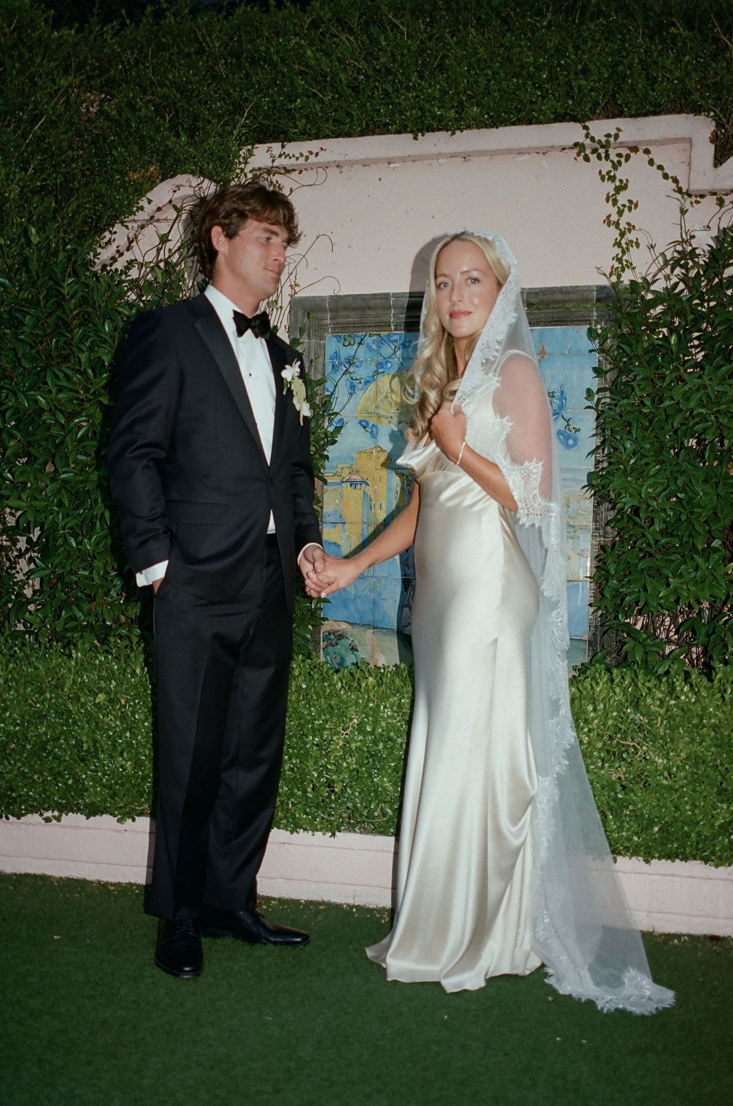 Wedding couple poses against a white wall with climbing vines and blue artwork.