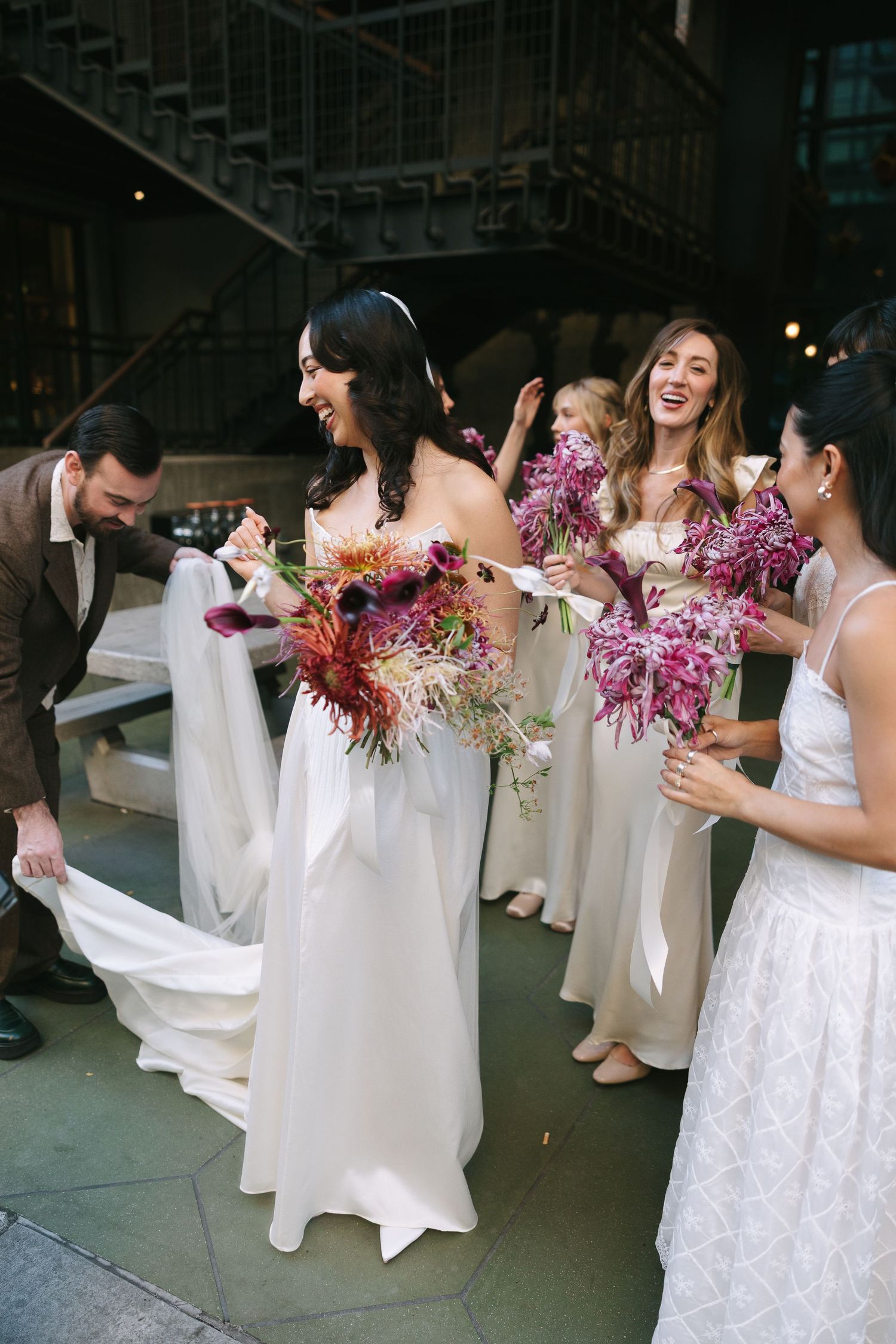 Bridesmaids in white dresses holding vibrant purple bouquets laugh and celebrate together at a wedding.