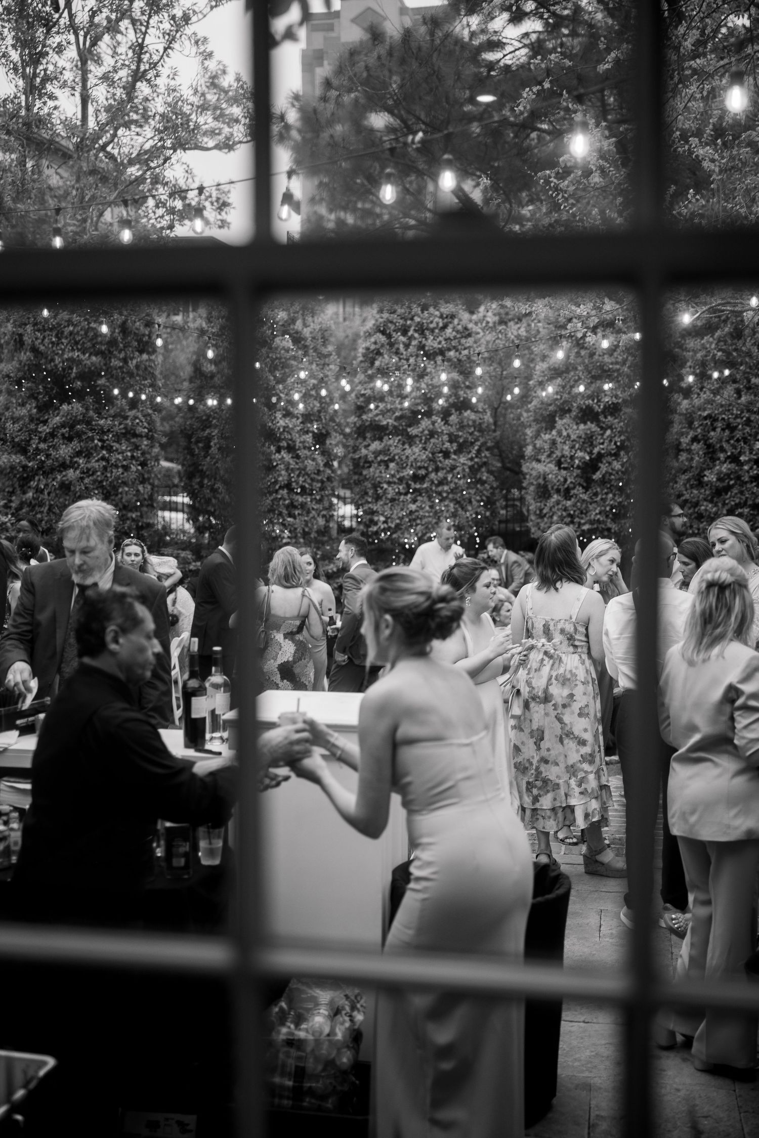 Black and white photo of wedding reception guests mingling under string lights at dusk.