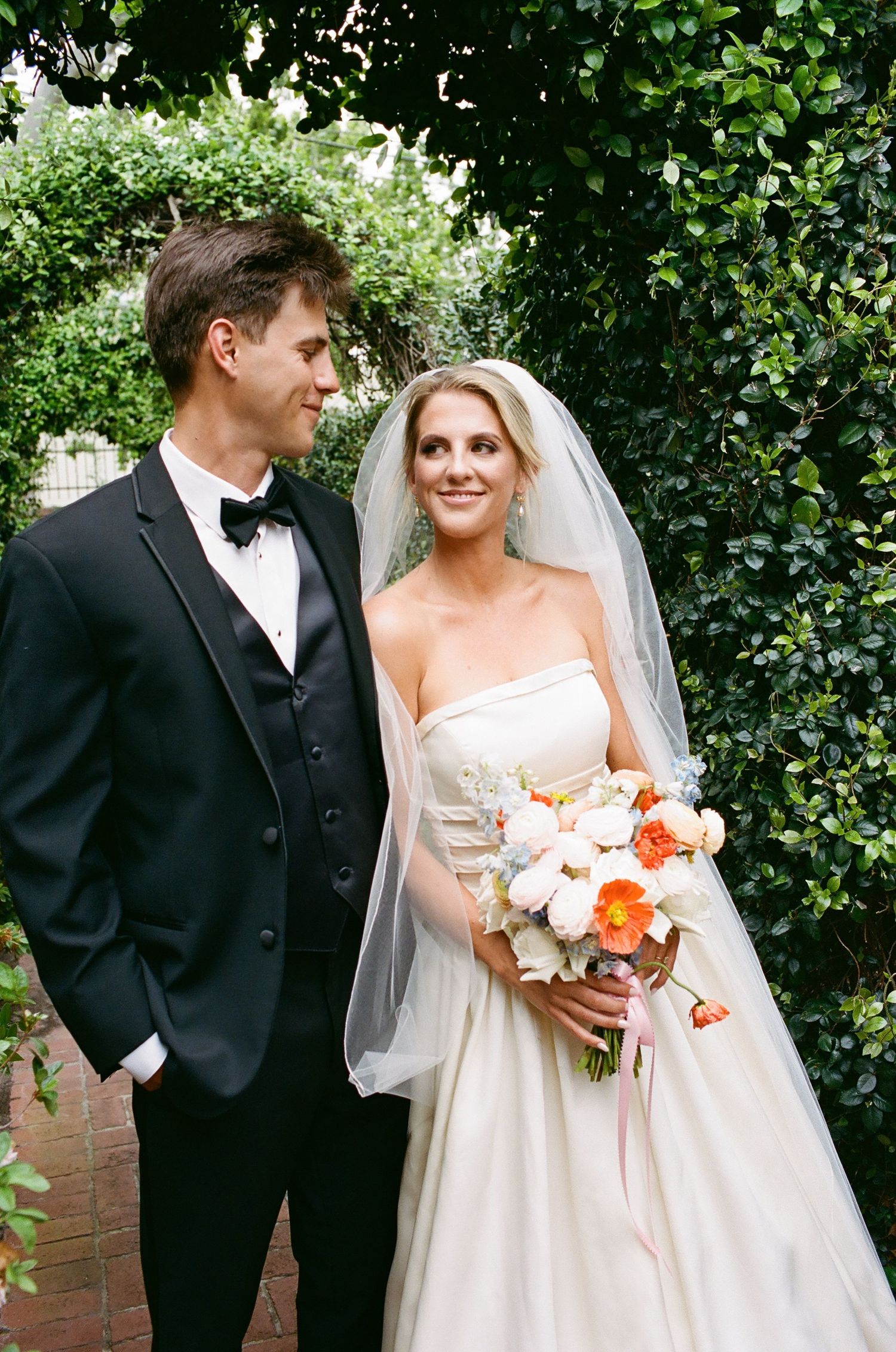 A couple poses against green foliage while holding a peach and white floral bouquet.