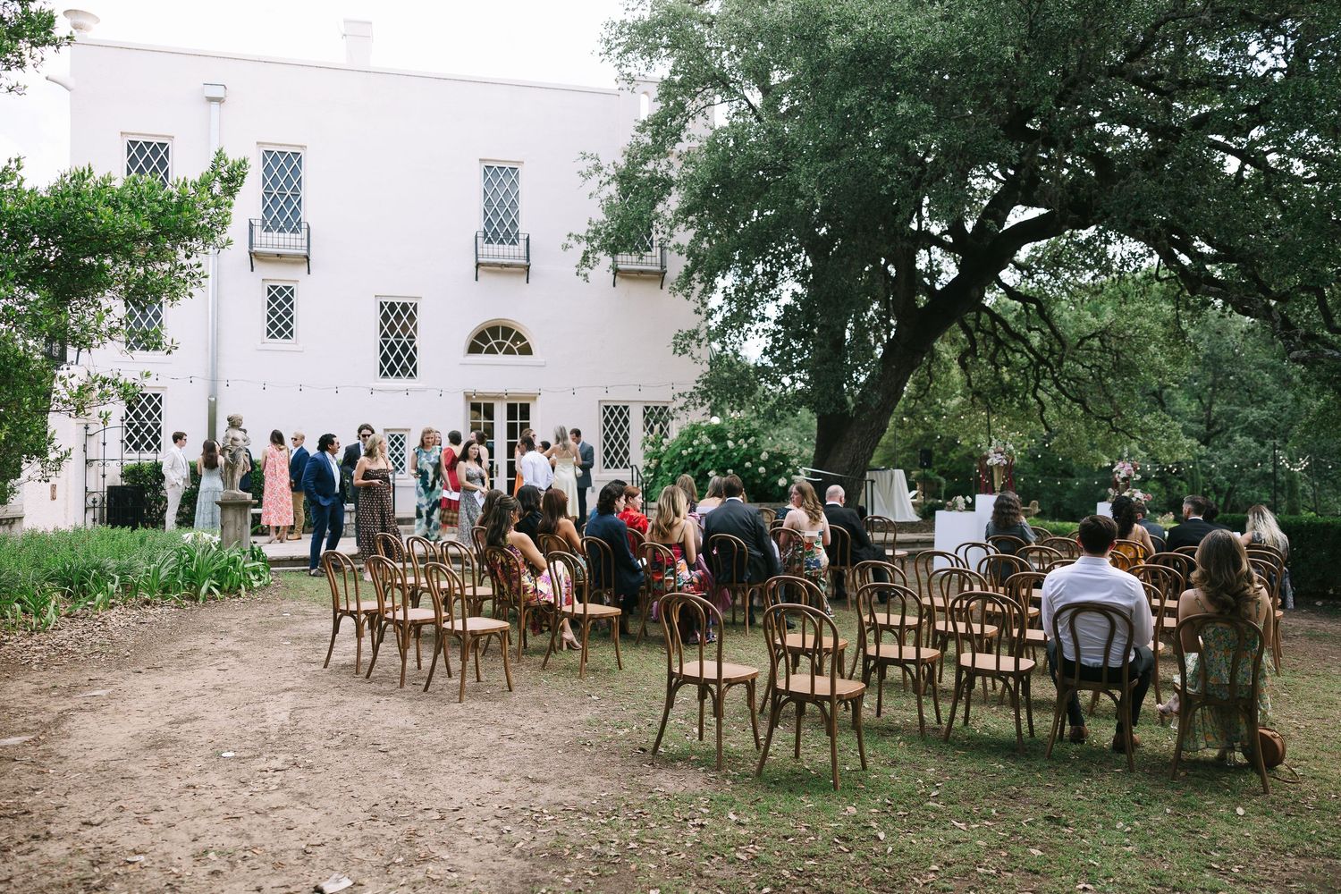 Wooden chairs arranged outdoors for a garden ceremony with guests gathering in the background.