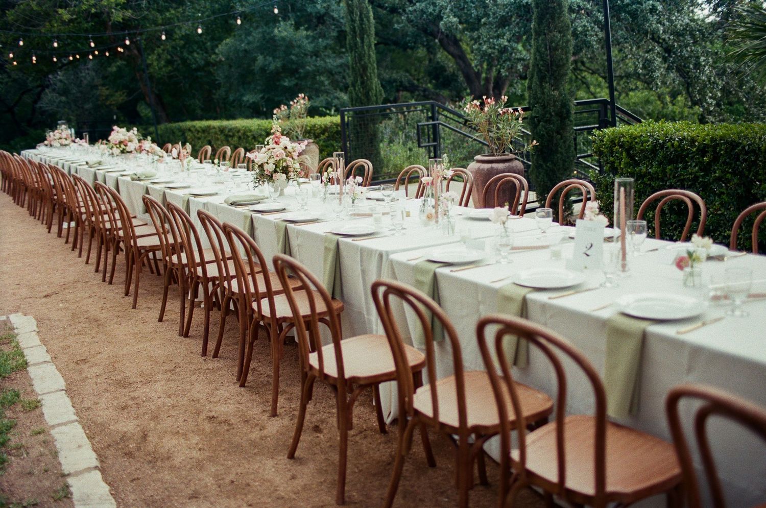 Long banquet table with white tablecloths and wooden chairs arranged outdoors for a garden wedding reception.