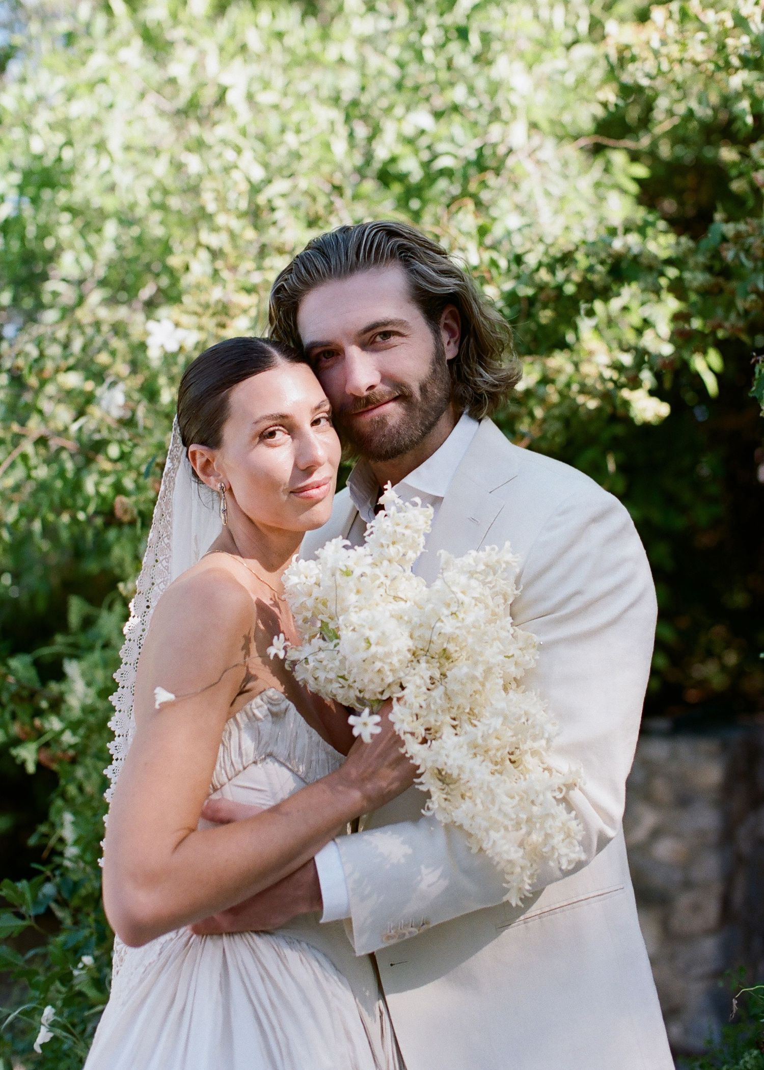 Bride and groom pose together holding white floral bouquet outdoors, wearing elegant ivory wedding attire among lush greenery.