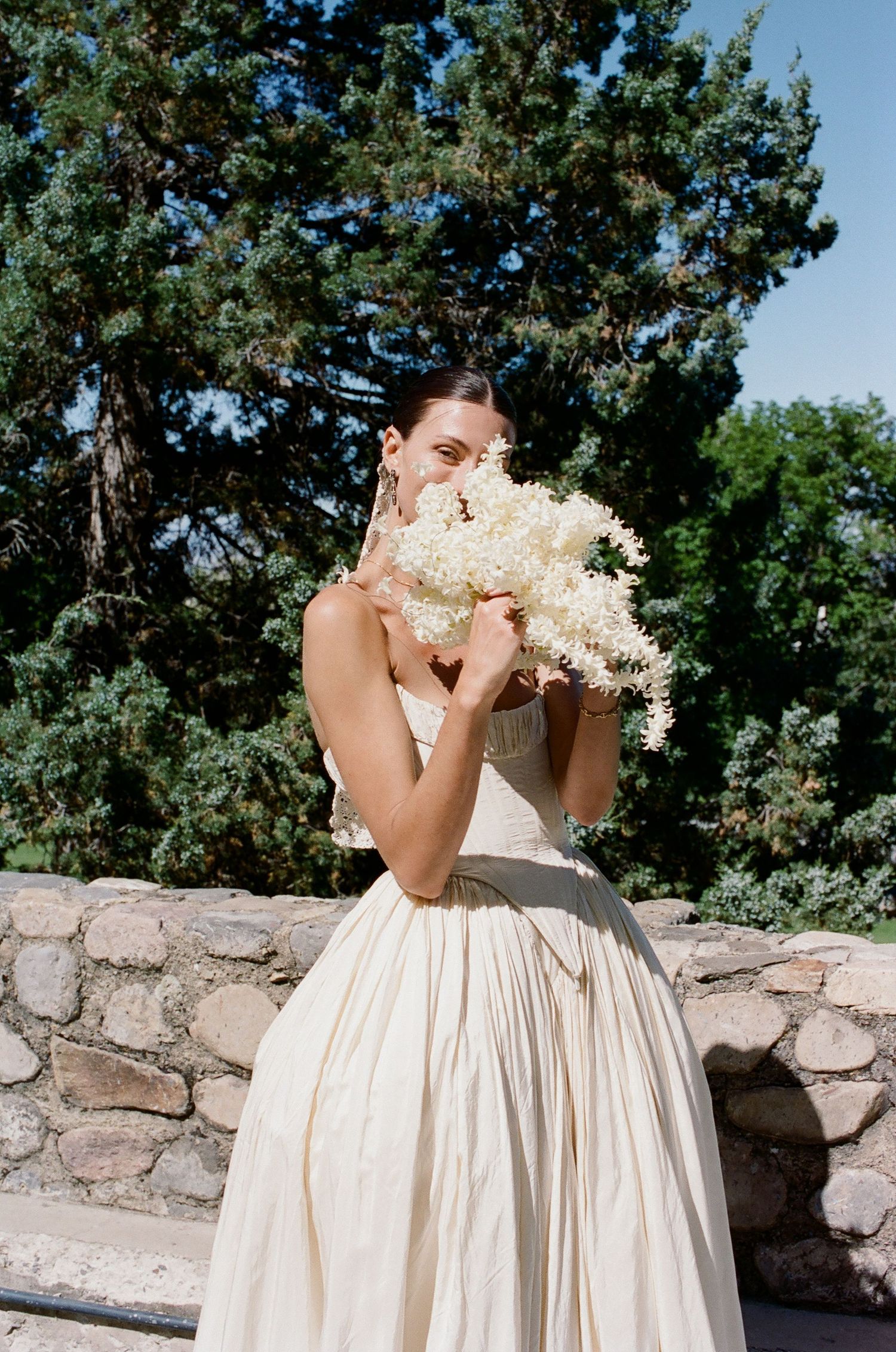 Bride in ivory ball gown smelling white floral bouquet outdoors with stone wall and green trees behind her.