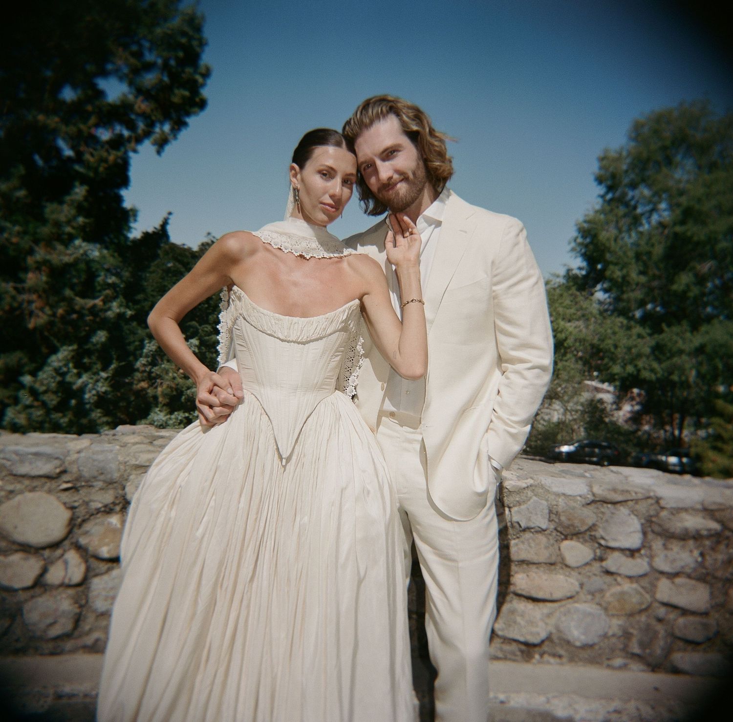 Elegant couple in white wedding attire posing outdoors near stone wall surrounded by lush green trees.