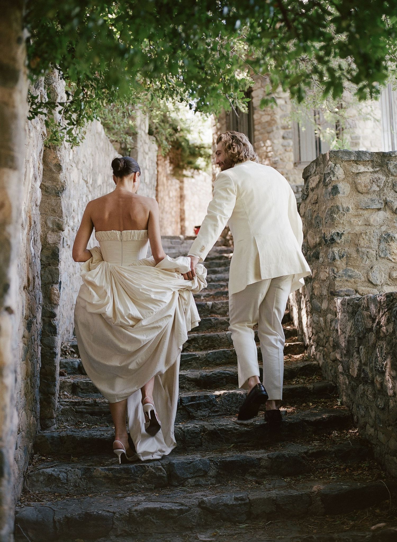A bride in a strapless gown and groom in a white suit climb stone steps at an outdoor wedding venue.